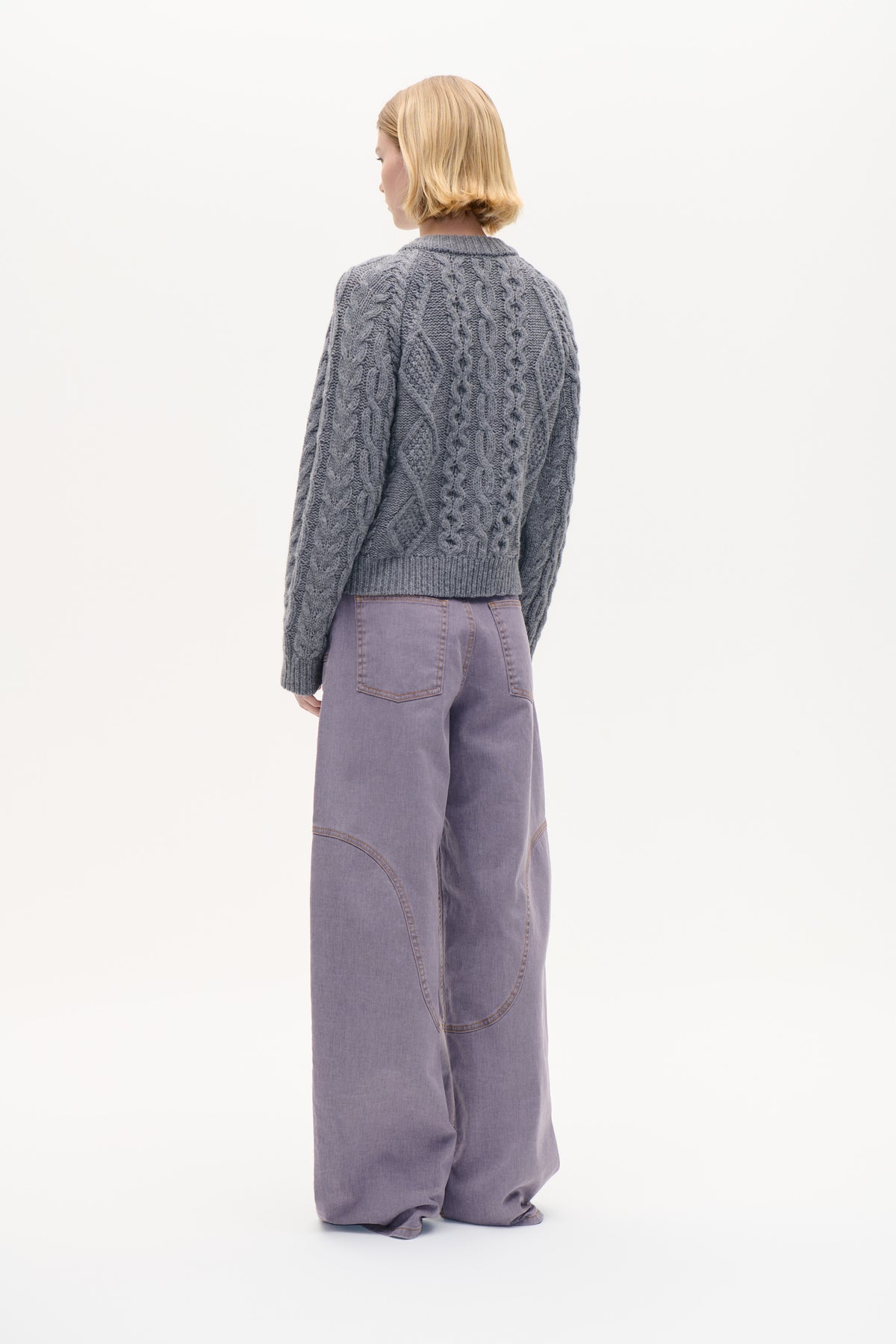 Back view of a model wearing a grey cable-knit sweater with ribbed trim and loose lavender jeans.