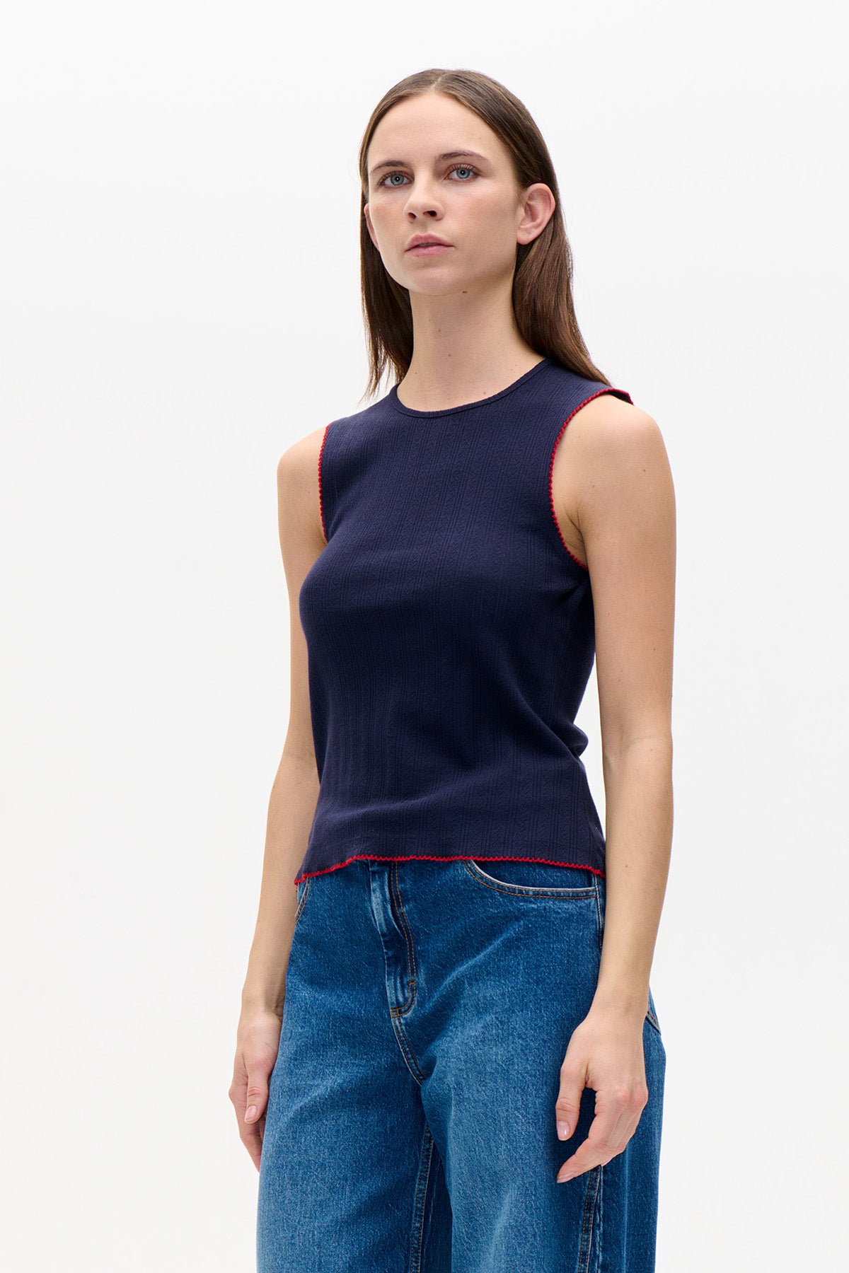 Le mannequin porte un haut sans manches bleu marine avec un subtil motif côtelé vertical et un bord festonné rouge, associé à un jean bleu.