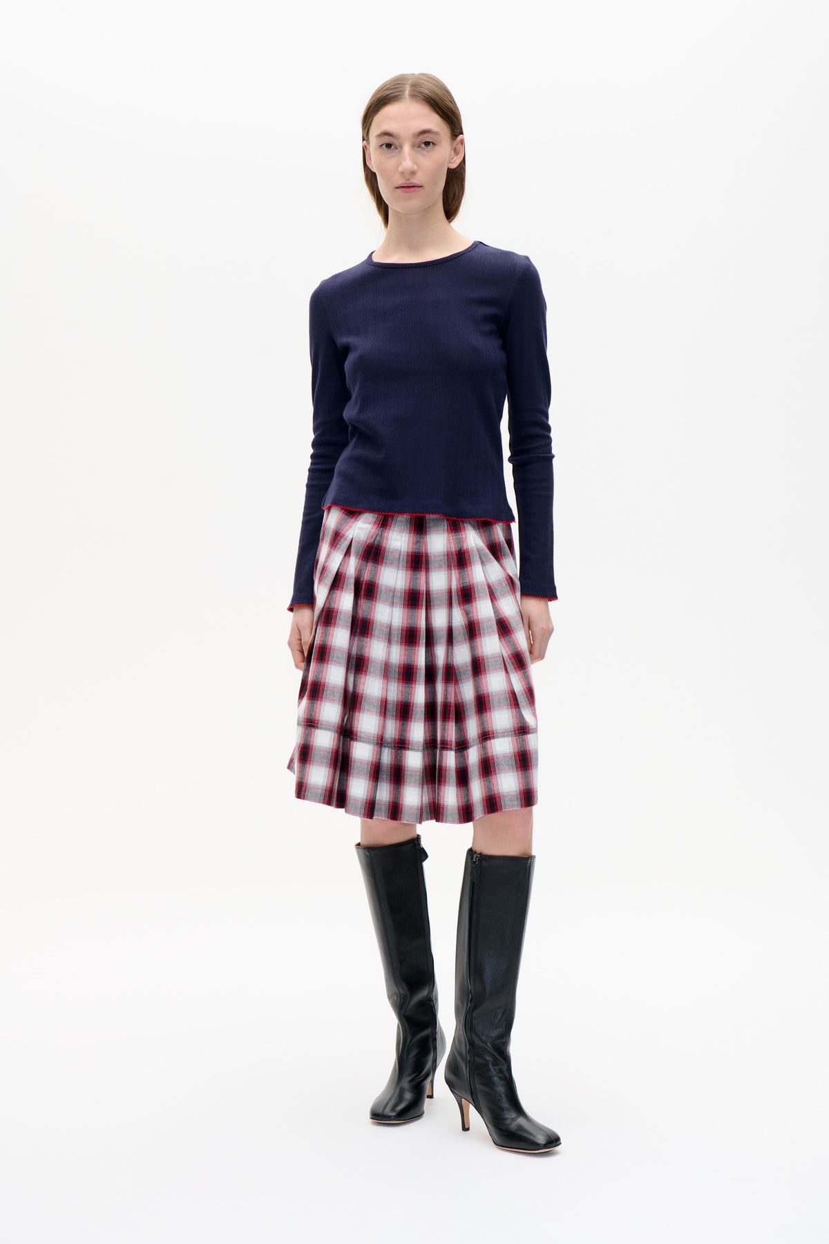 Mannequin portant un pull en maille bleu marine à manches longues, à texture verticale discrète et bordure rouge, et une jupe plissée à carreaux rouge, blanc et gris.
