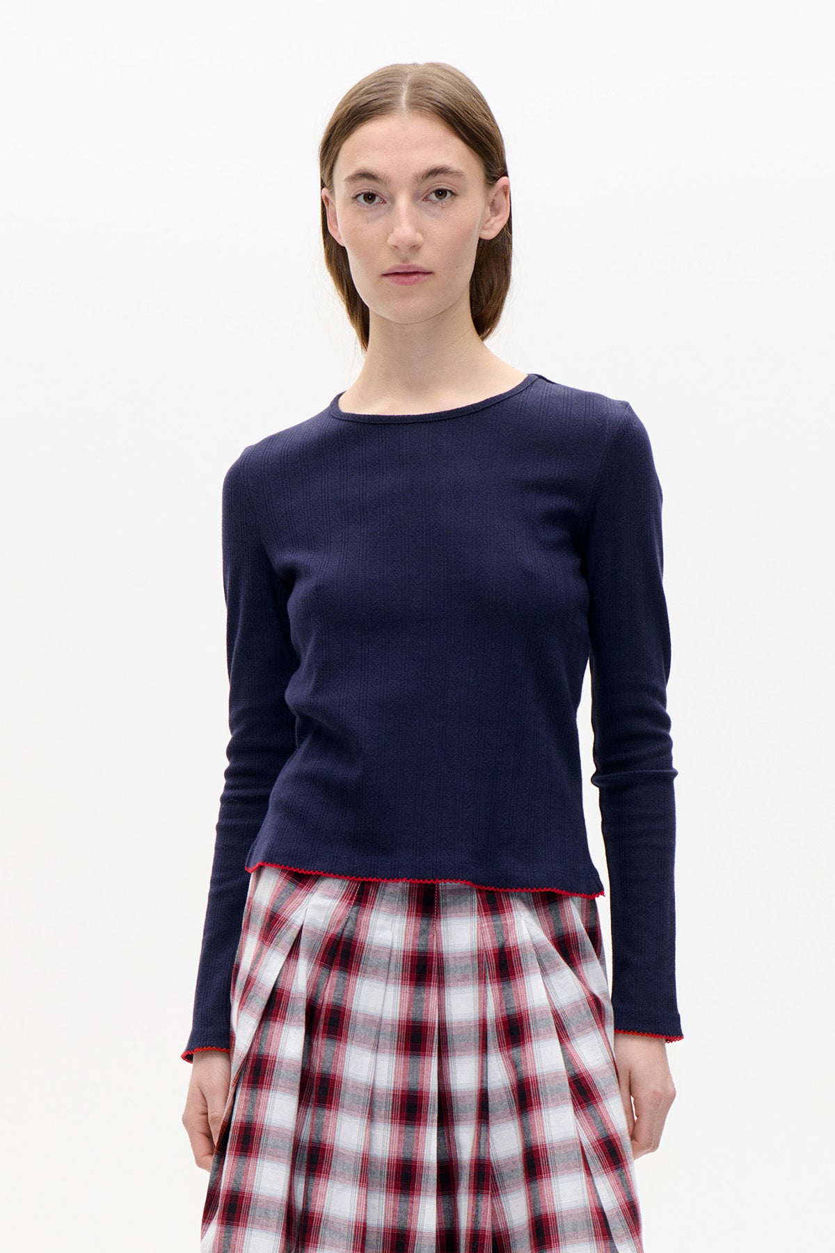 Vue de face d'un mannequin portant un haut bleu marine à manches longues, à une légère texture verticale et une bordure picot rouge, associé à une jupe à carreaux rouge, blanc et gris.
