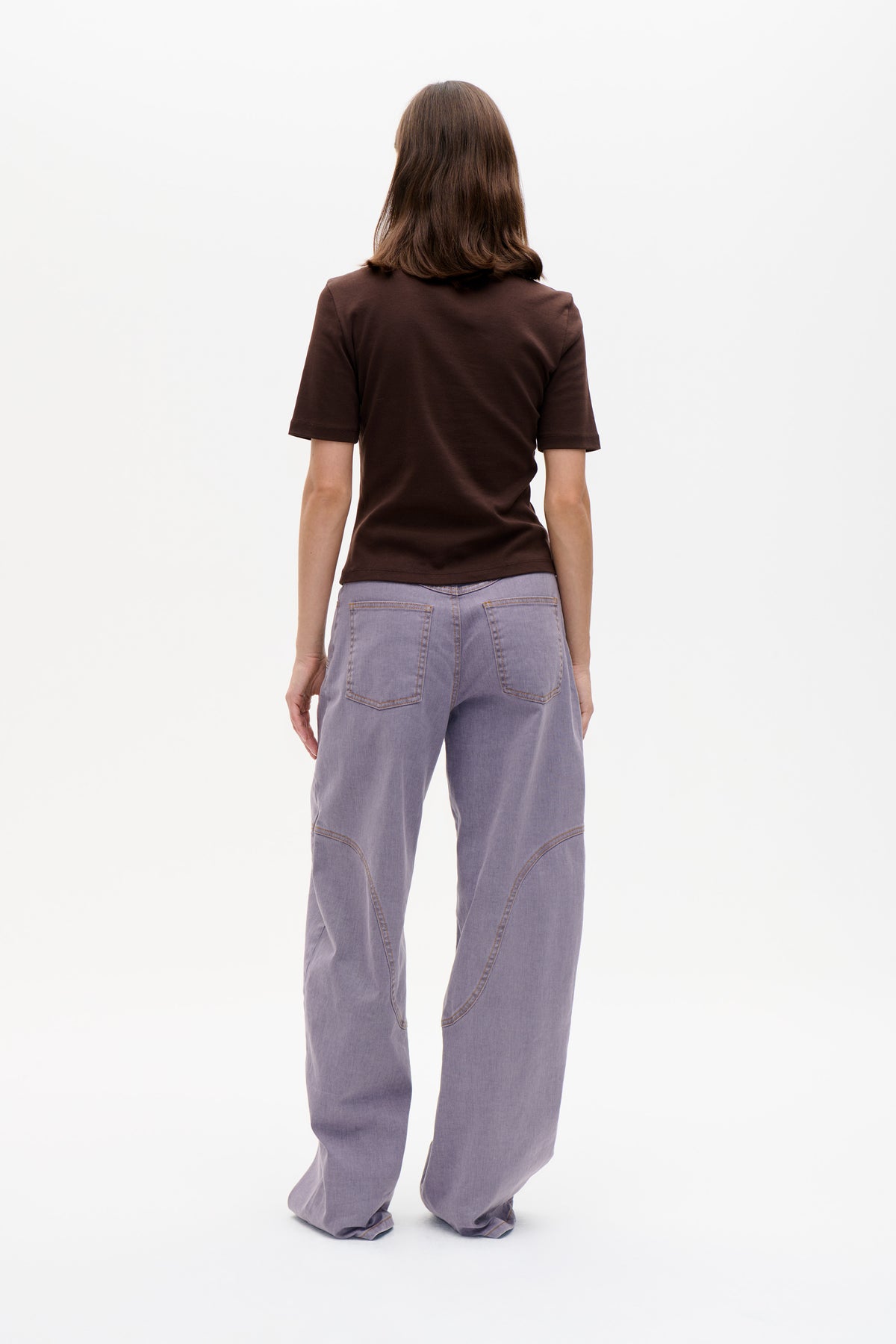 Rückenansicht eines Models, das ein schlichtes dunkelbraunes T-Shirt mit kurzen Ärmeln und eine locker sitzende lavendelfarbene Jeans mit geschwungenen Nahtdetails trägt.