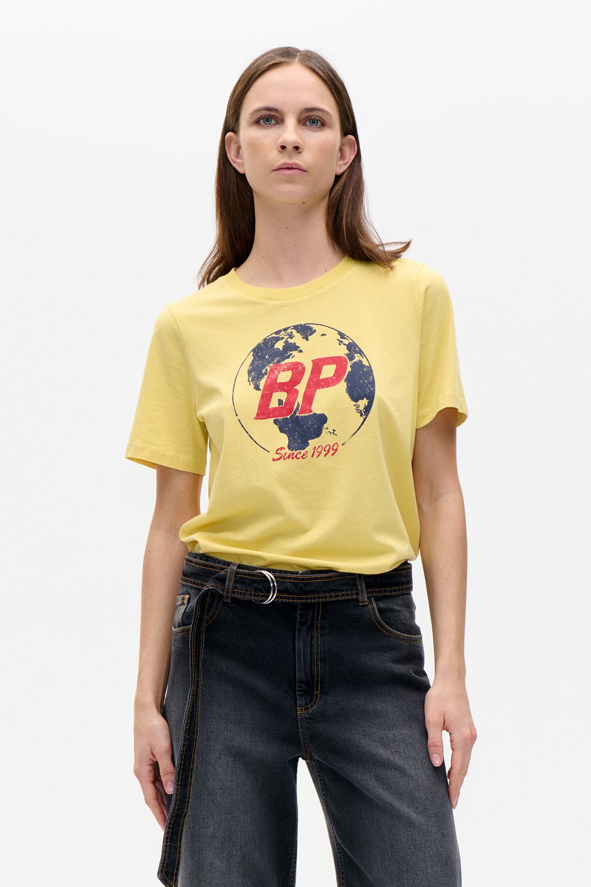 Model wearing a pale yellow short-sleeved T-shirt with a globe and red BP graphic, paired with dark jeans.
