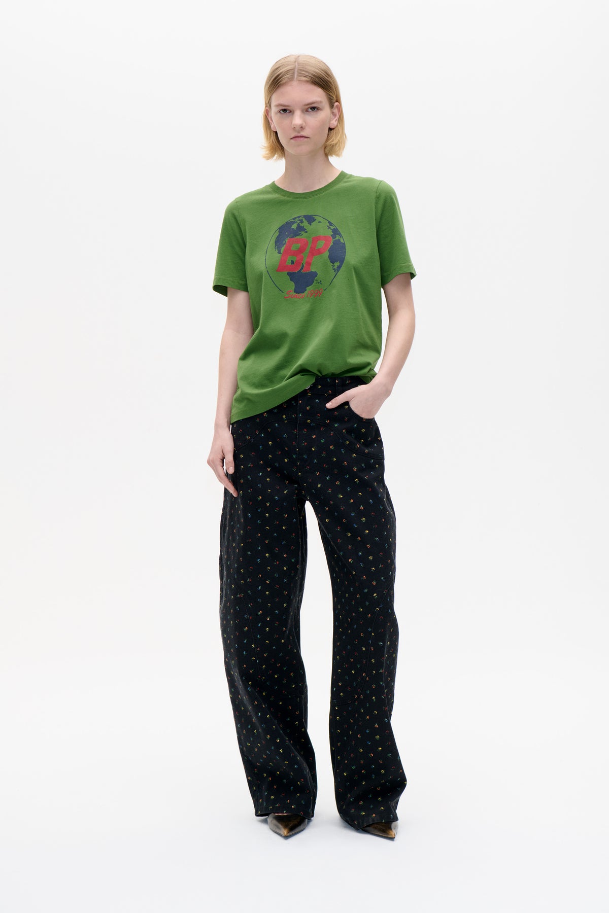 Front view of a model wearing a green short-sleeved T-shirt with a globe graphic and red “BP Since 1999” lettering, paired with black wide-leg trousers in a tiny multicolour print.