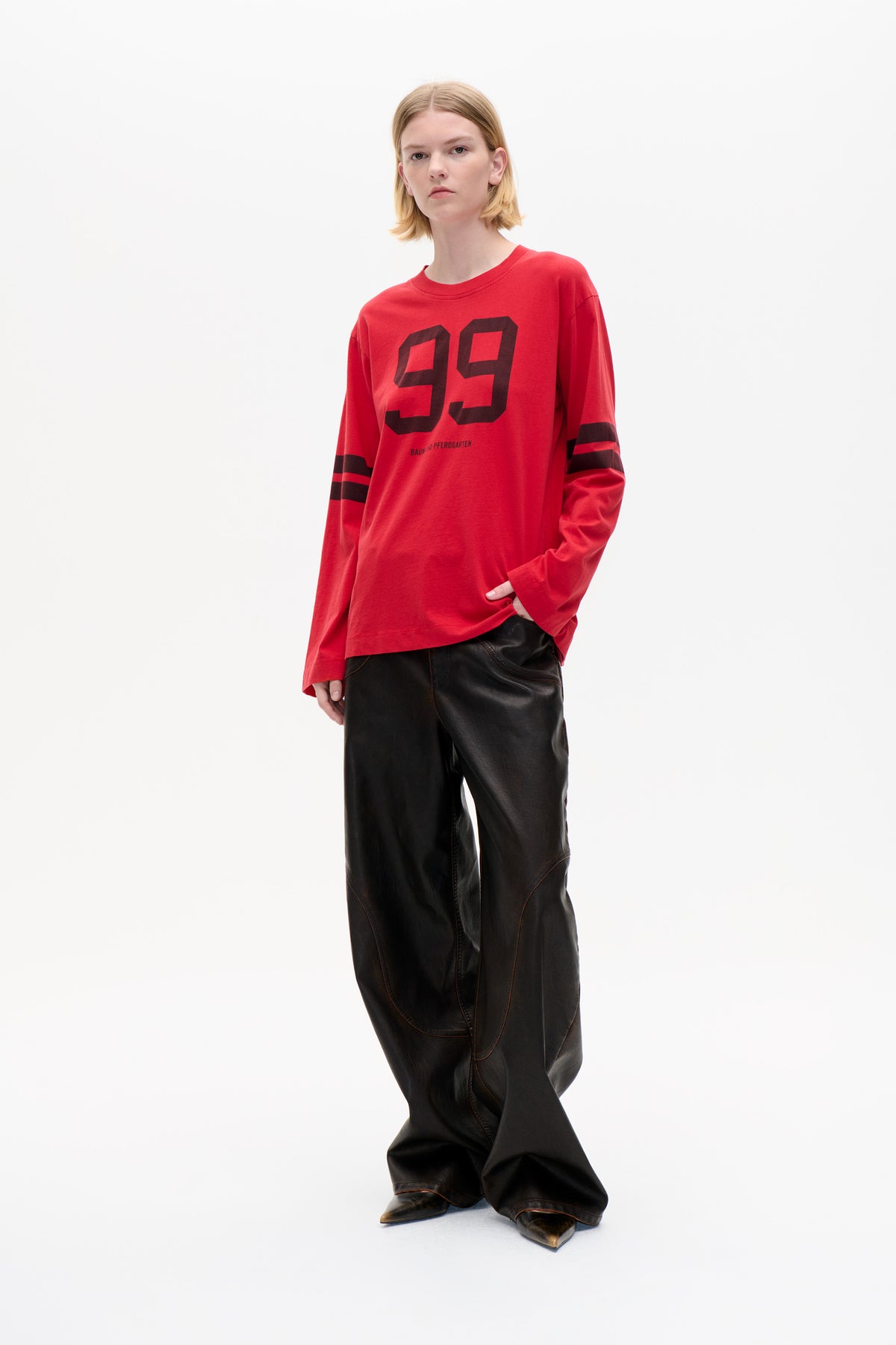 Model wearing a red long-sleeve T-shirt with a large black “99” print and sleeve stripes, paired with black trousers.
