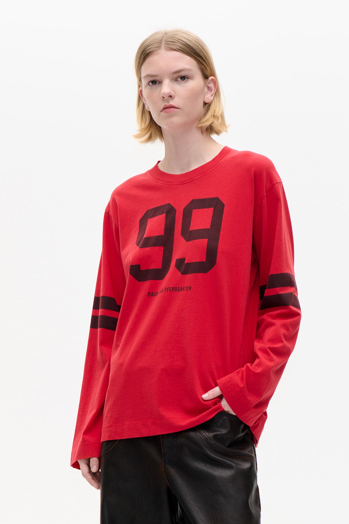 Model wearing a red long-sleeved T-shirt with a black number 99 graphic and sleeve stripes, paired with black trousers.