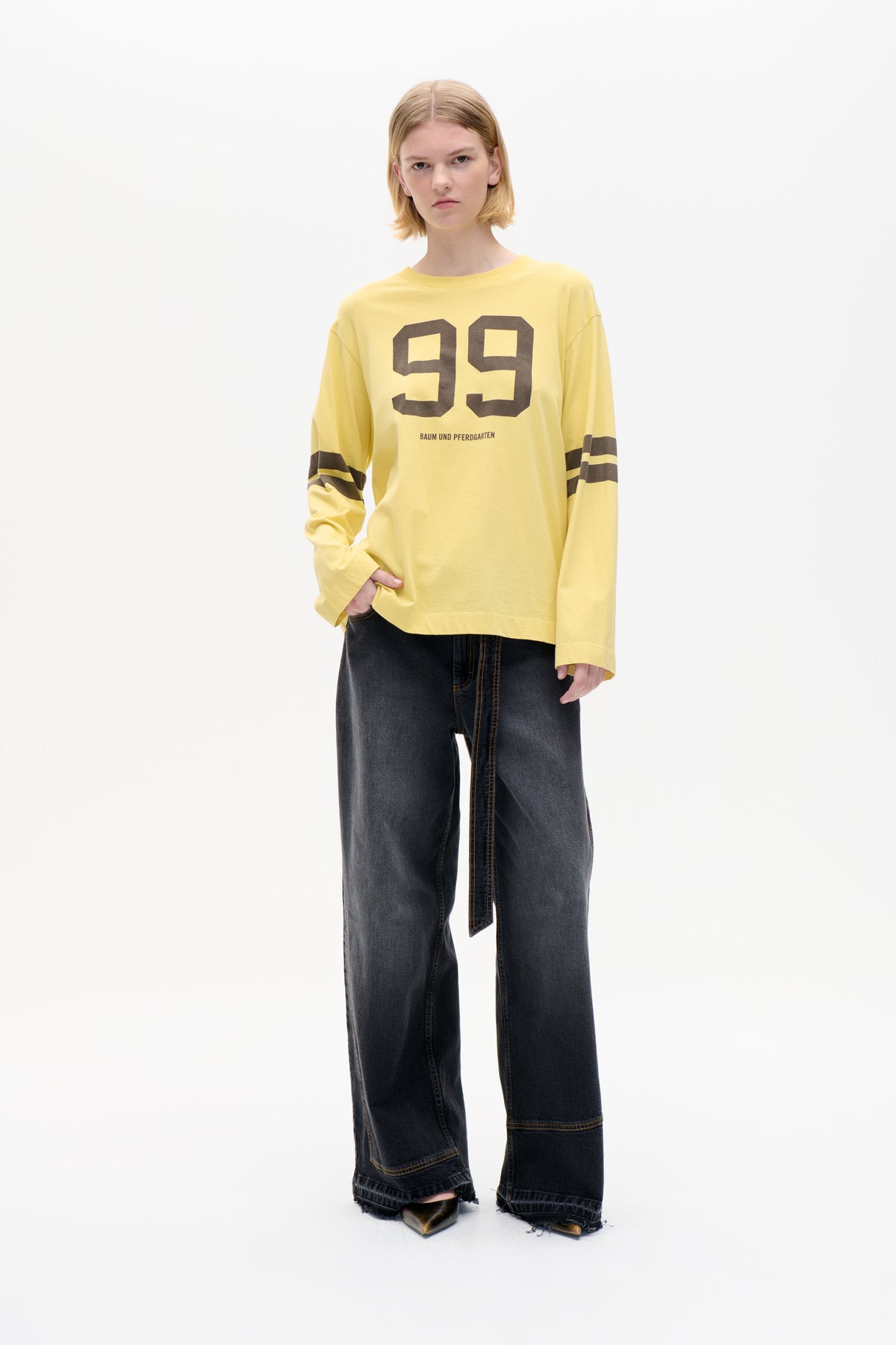 Model wearing a pale yellow long-sleeve T-shirt with a large dark 99 graphic and striped sleeves, paired with wide black jeans.