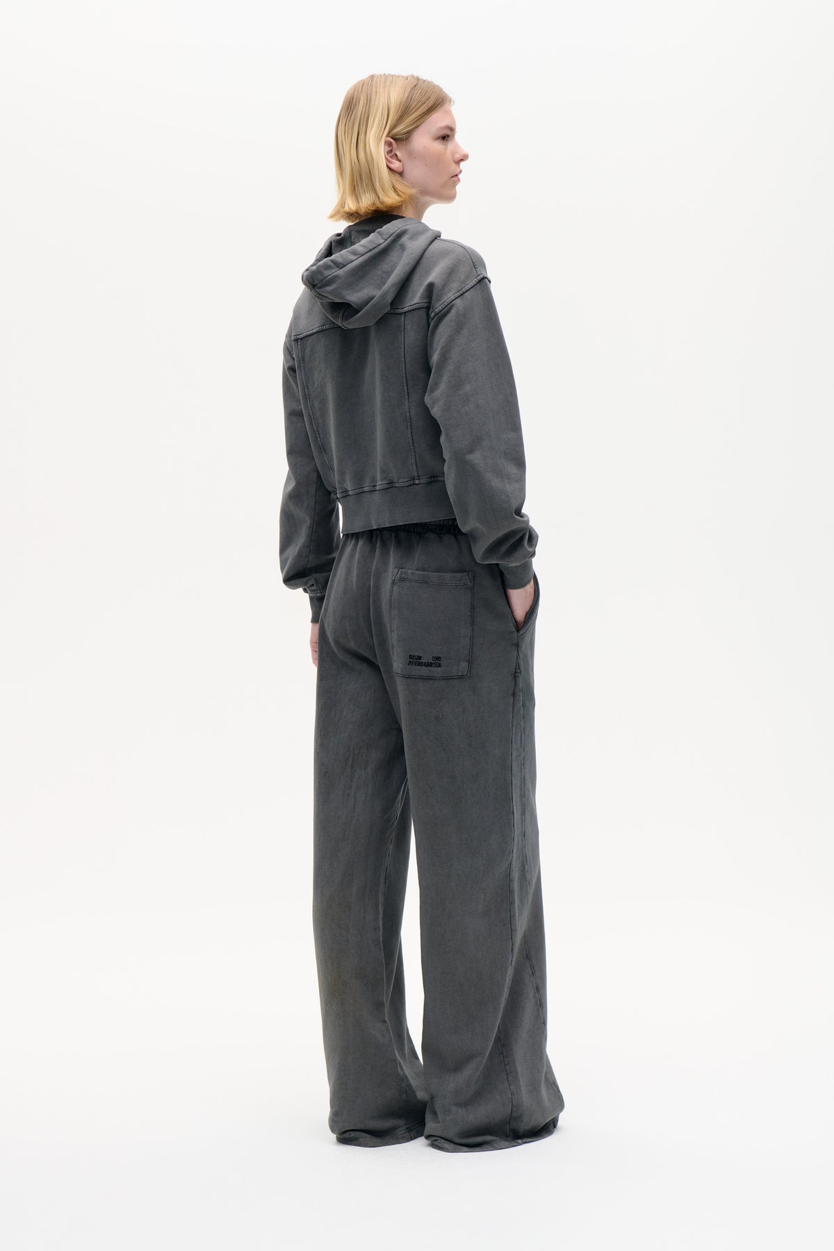 Model in a charcoal washed hoodie and matching wide-leg sweatpants, shown from behind with a hood and logo patch on the back pocket.