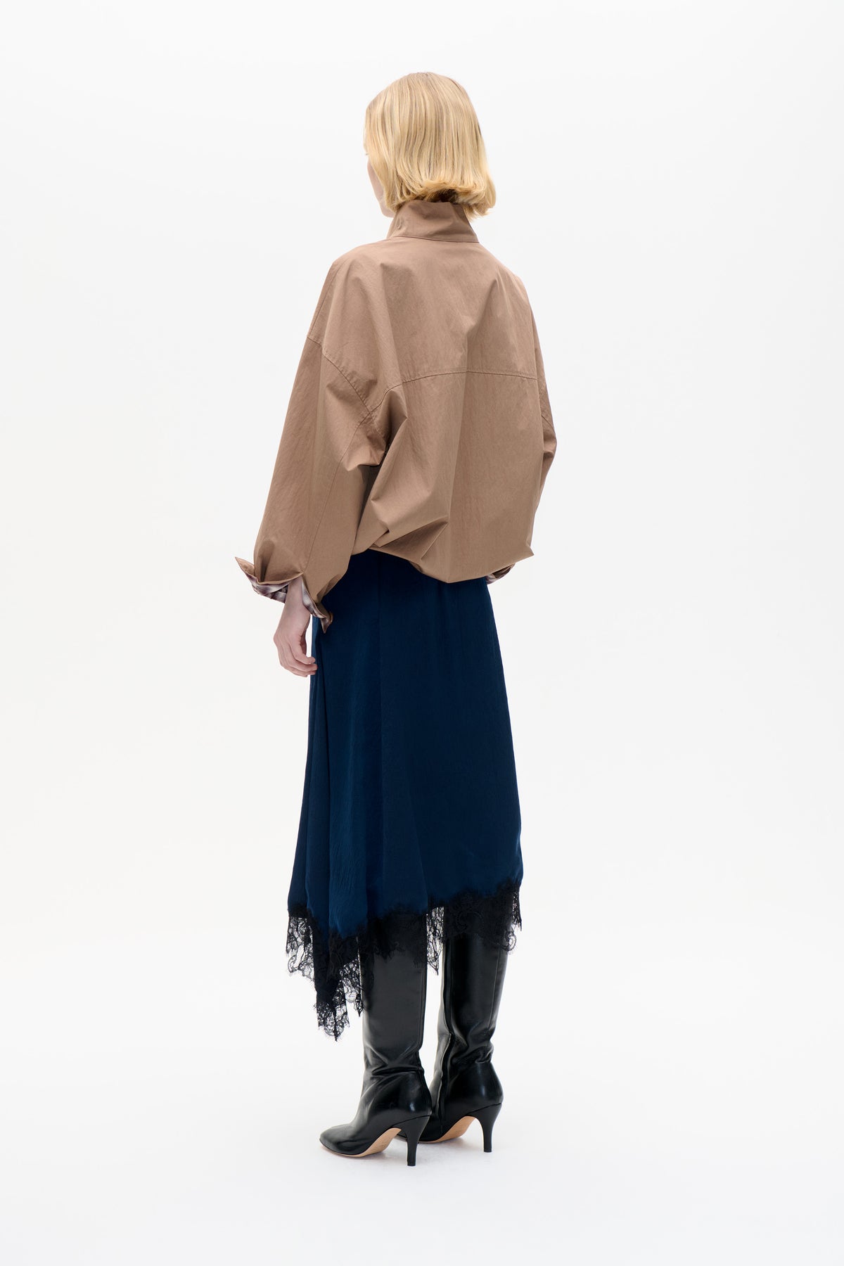Back view of a model wearing an oversized taupe jacket with a raised collar over a navy lace-trimmed skirt and black heeled boots.