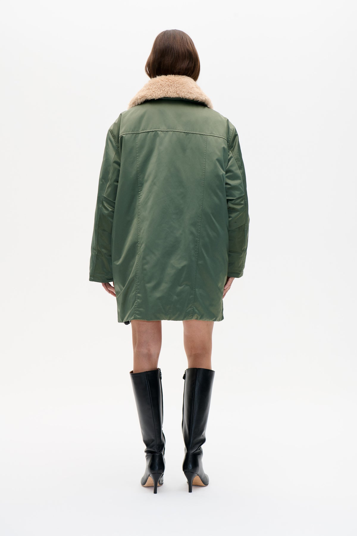 Back view of a model wearing a dark olive-green coat with a wide beige shearling collar and stitched back panel.