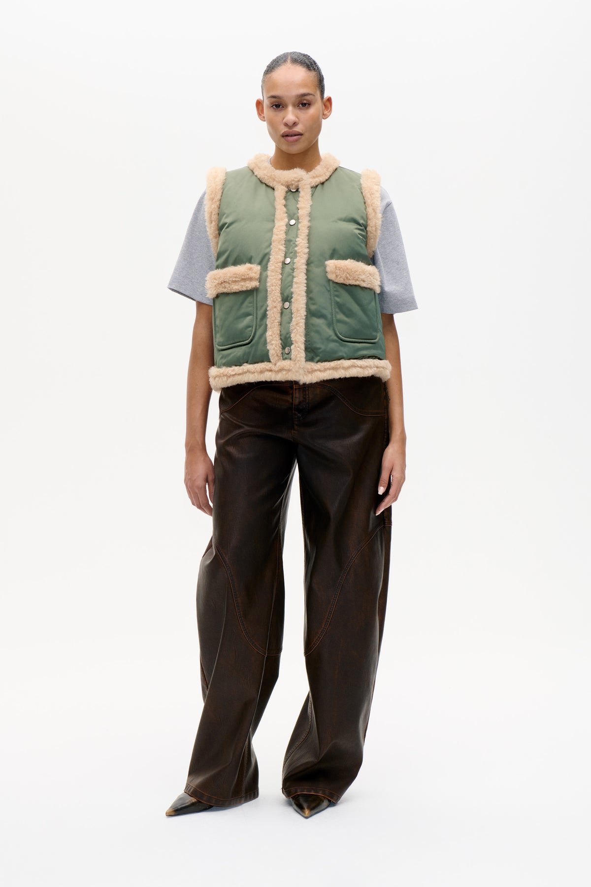 Model wearing a sage-green sleeveless vest with cream fluffy trim and snap buttons over a grey T-shirt, paired with dark brown wide-leg trousers.