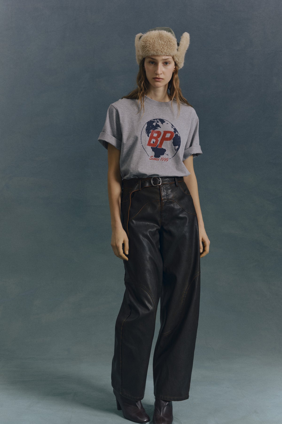 Model against a blue-grey backdrop wearing a cream fuzzy hat, grey globe-print T-shirt marked “BP Since 1999,” and black wide-leg trousers.