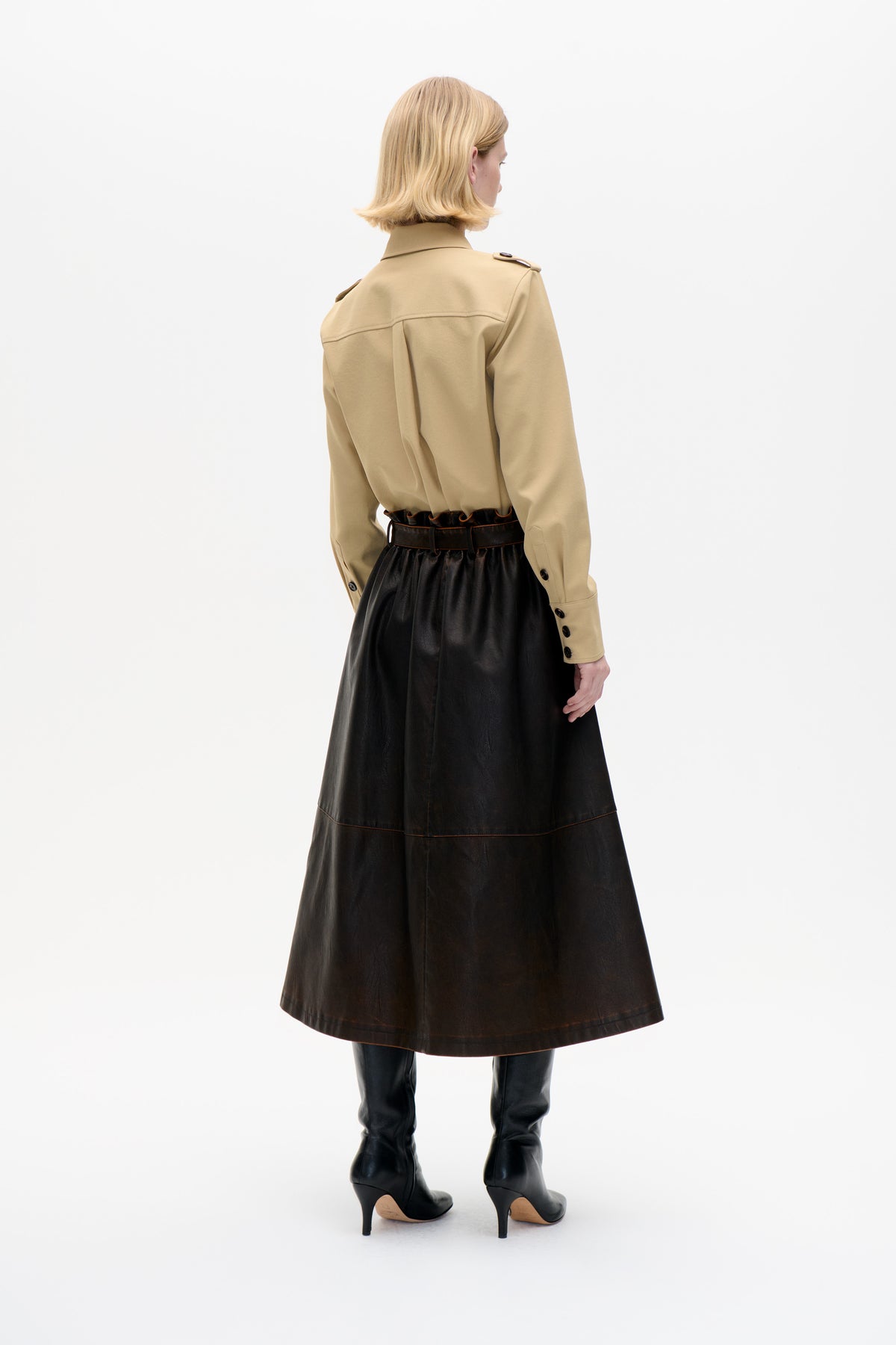 Rear view of a model wearing a sand-beige collared jacket with shoulder tabs and button cuffs, paired with a black flared skirt and tall boots.