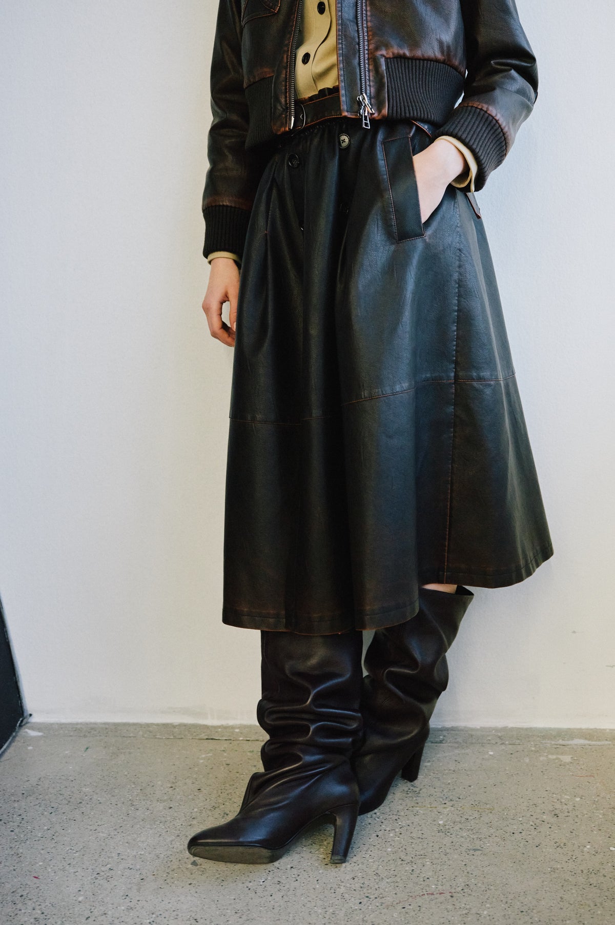 Cropped campaign view of a model wearing a dark leather-look jacket over a long black paneled skirt and trousers, with black heeled boots.