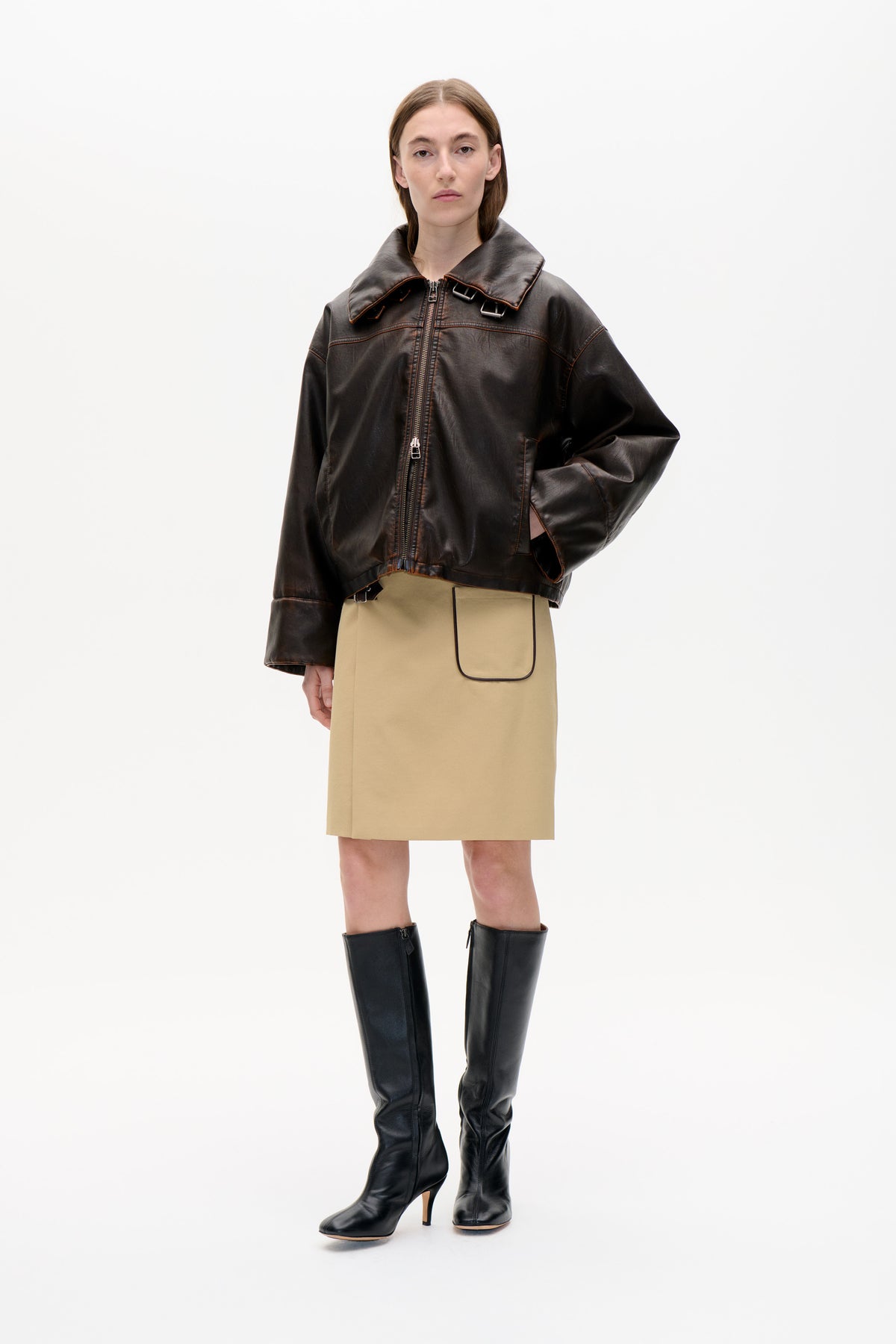 Front view of a model wearing a glossy dark brown oversized zip-front jacket with a wide buckled collar, beige skirt and black knee-high boots.
