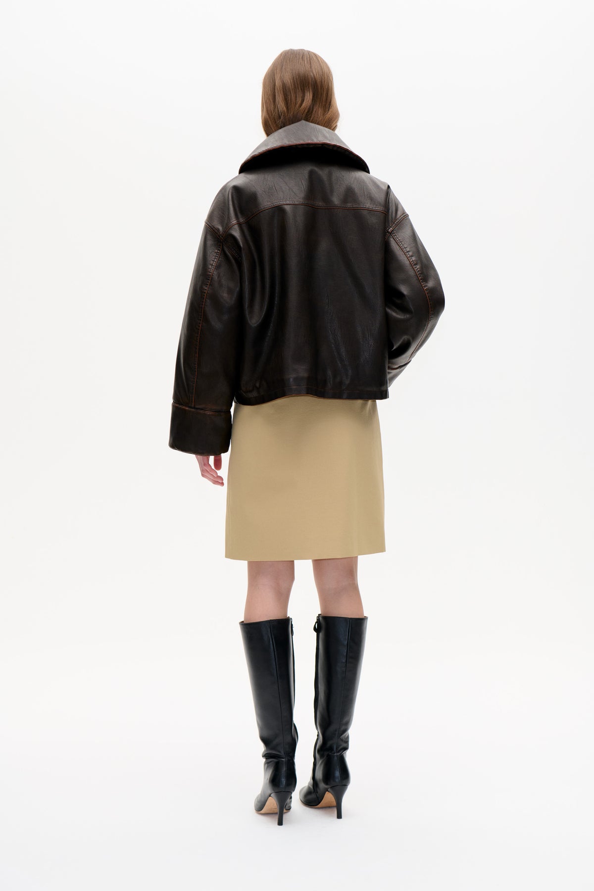 Back view of a model wearing a dark brown leather-look jacket with a wide collar and contrast stitching, paired with a beige skirt.