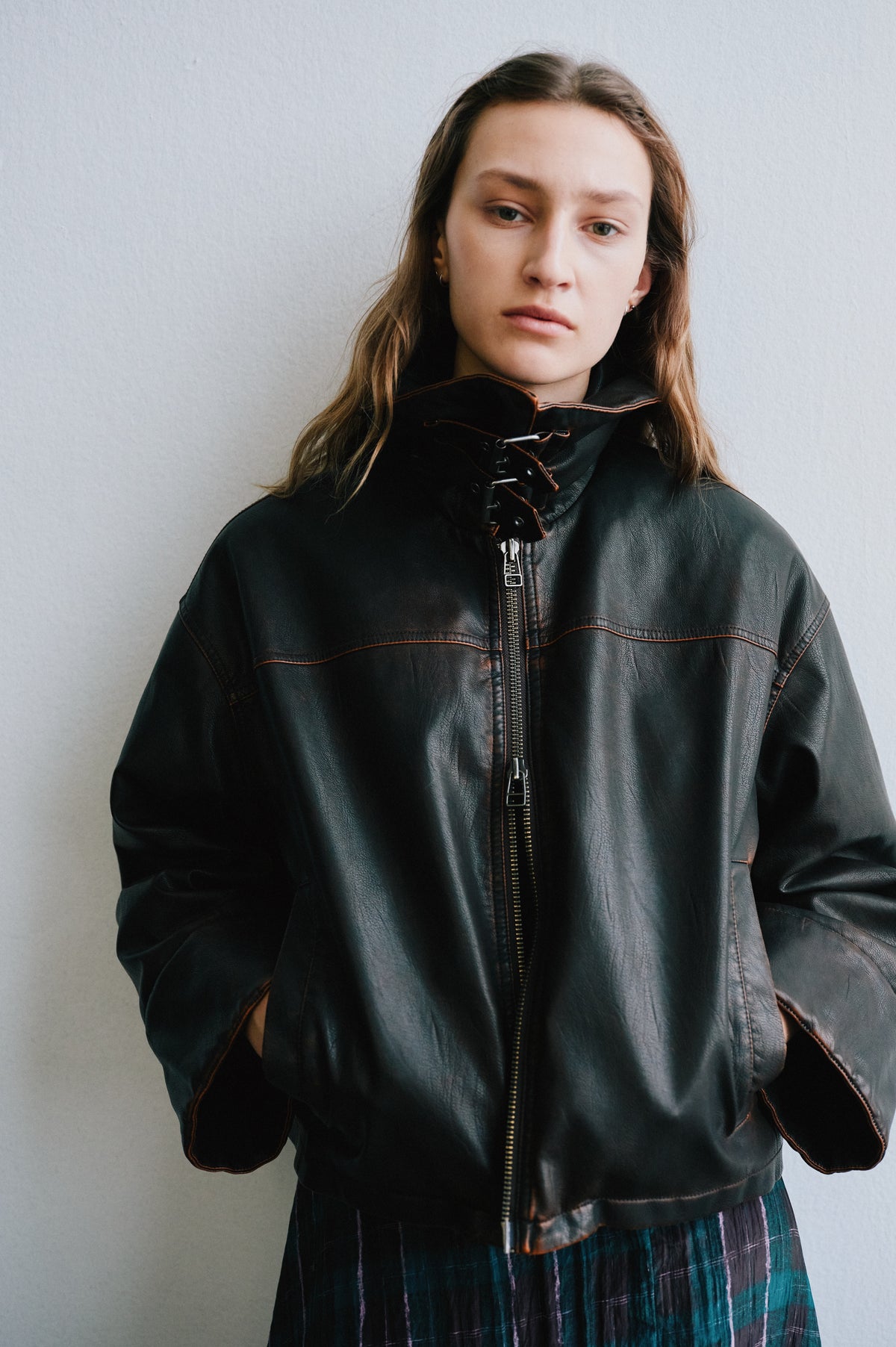 Campaign portrait of a model in a black leather-look zip jacket with brown contrast stitching and a teal and purple plaid skirt.
