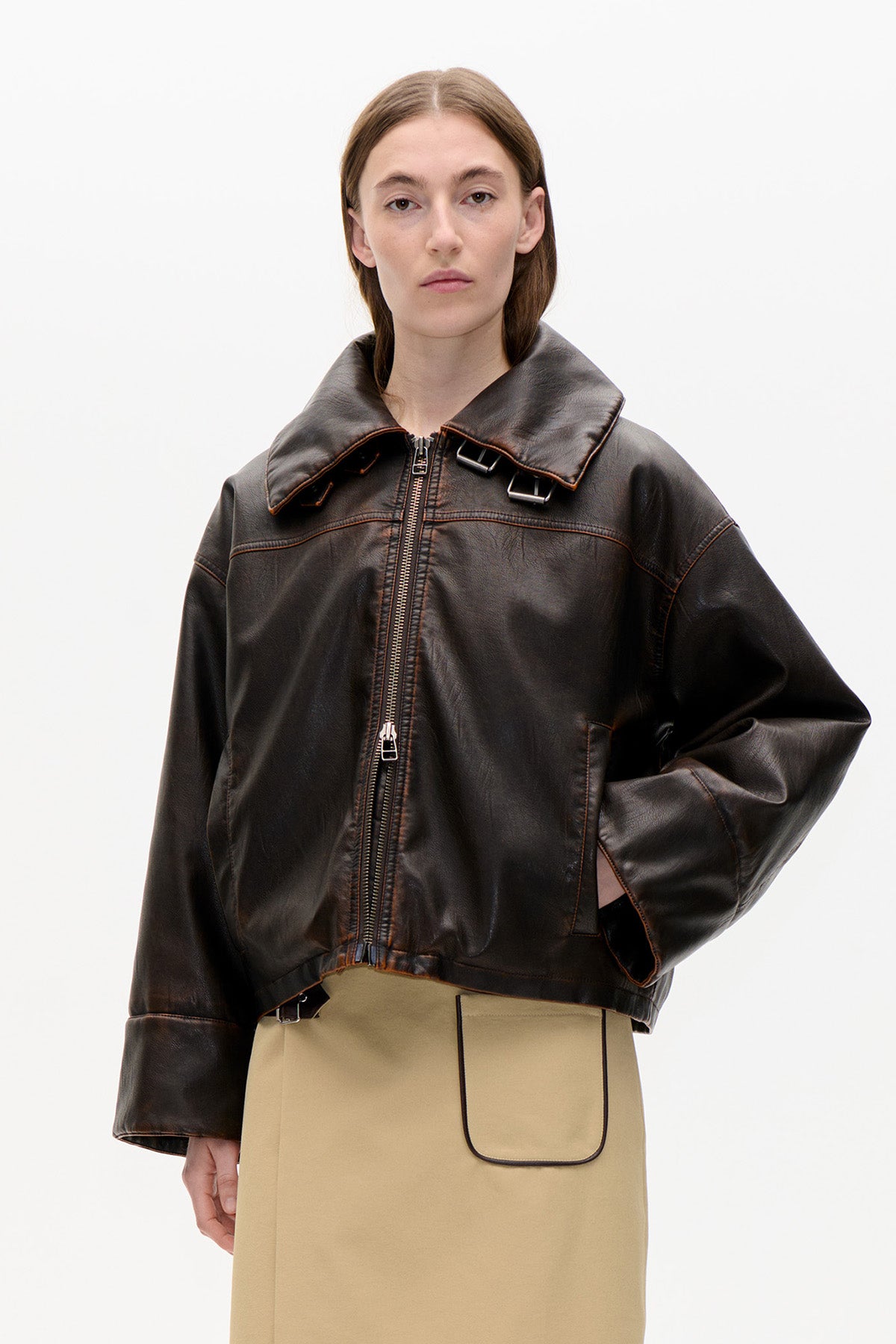 Front view of a model wearing a dark brown oversized jacket with a wide collar, zip fastening and buckle details, over a beige skirt.