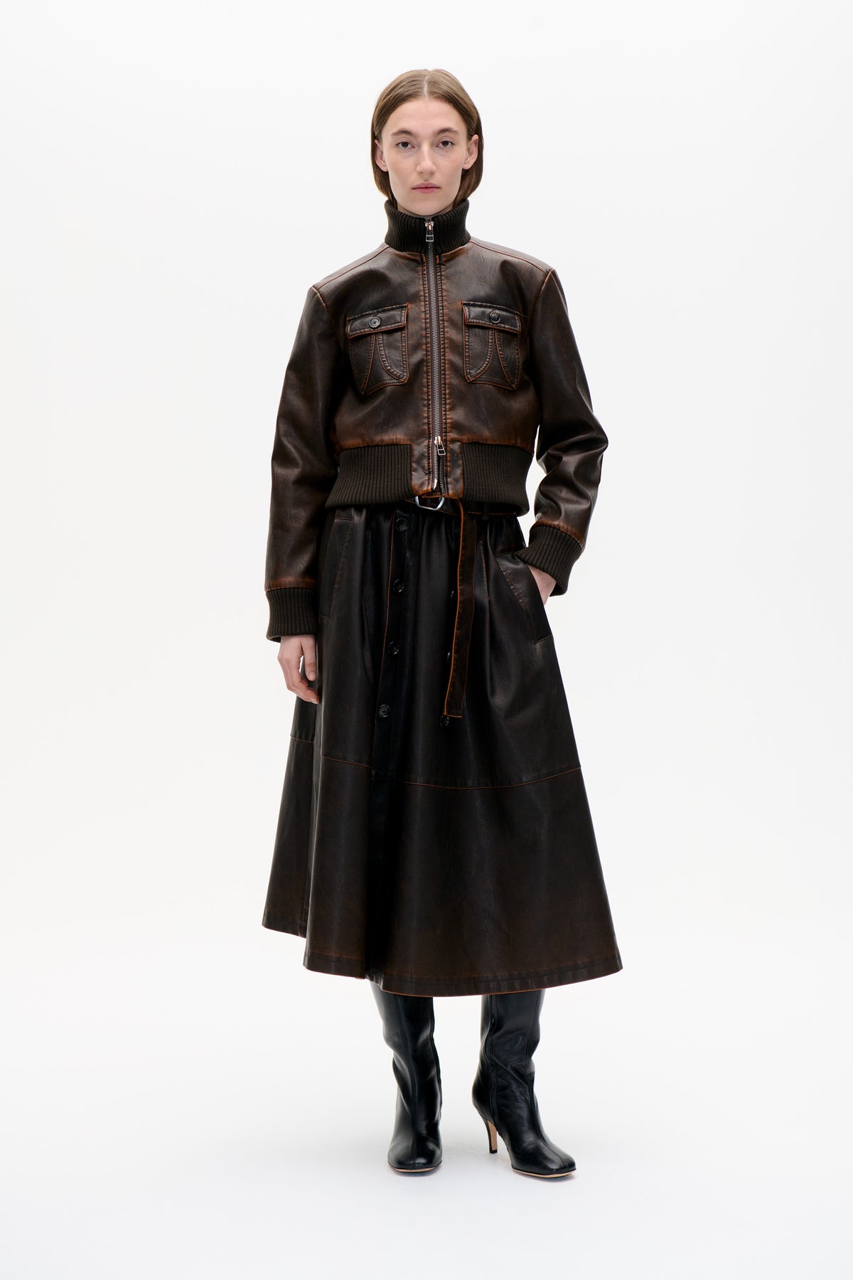 Front view of a model wearing a dark brown zip-front jacket with ribbed black trim and flap pockets, over a black button-front skirt.