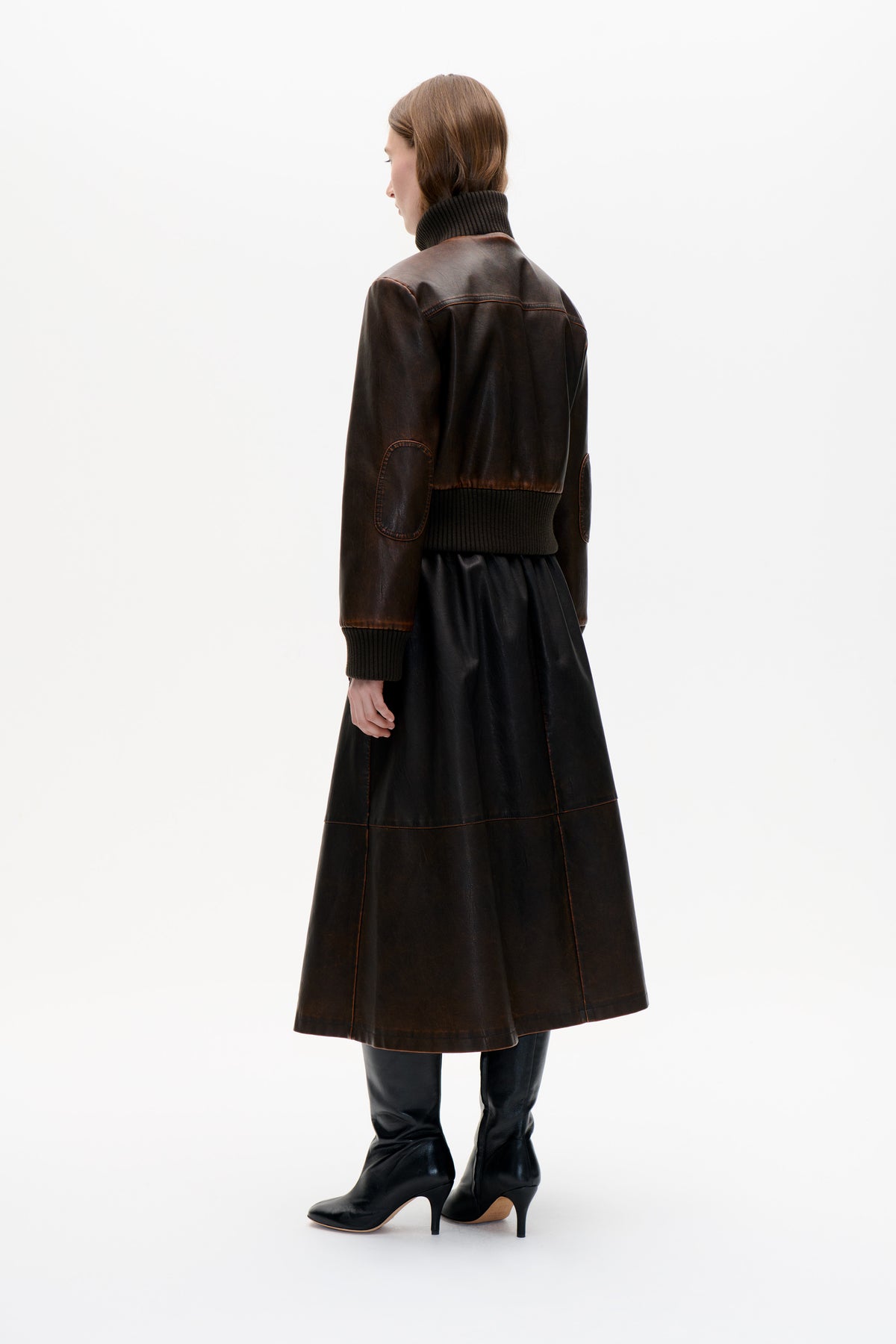 Model seen from behind in a dark brown leather-look jacket with ribbed trim and elbow patches, paired with a long black skirt.