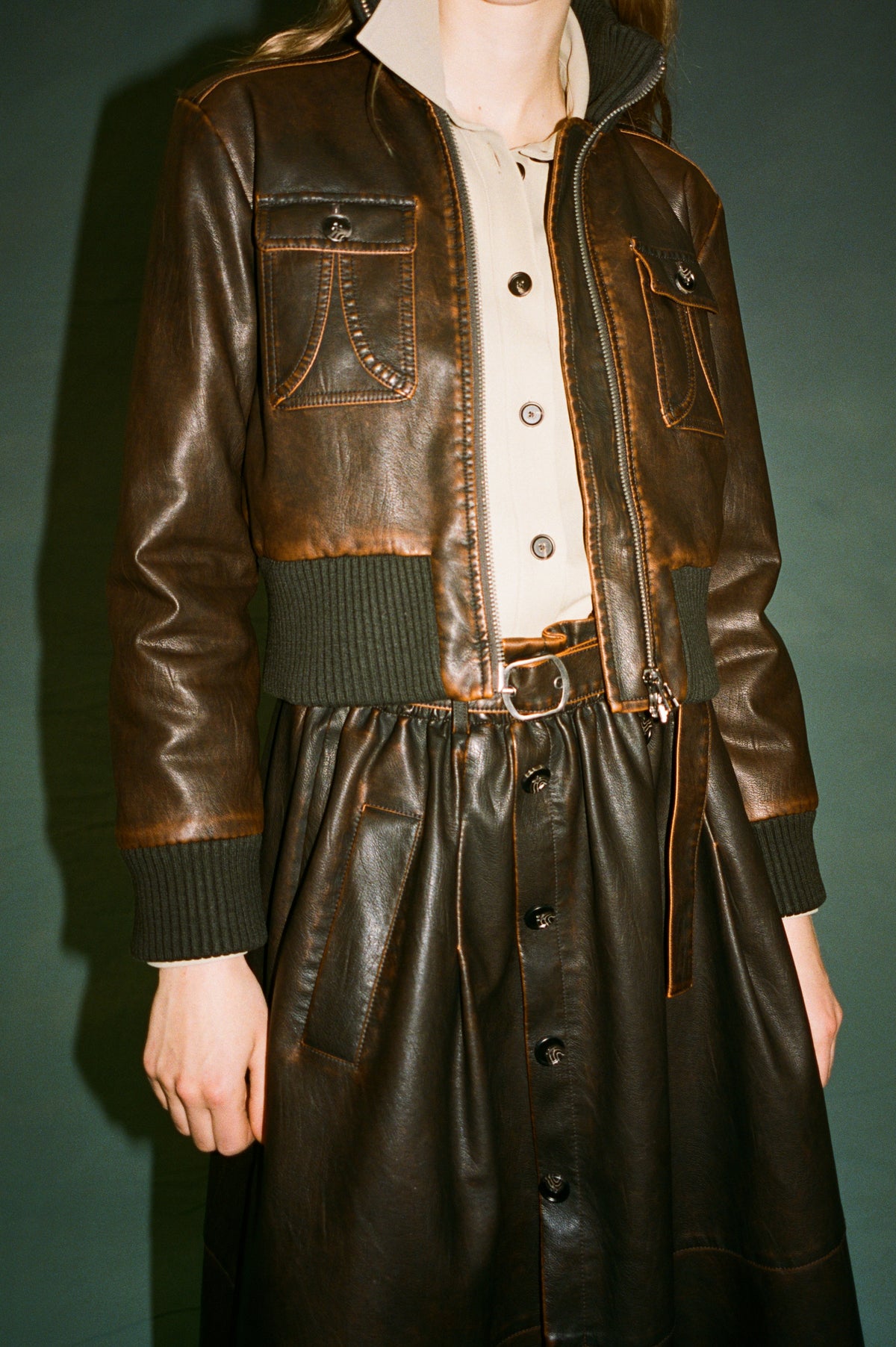 Campaign close-up of a model wearing a brown leather-look zip jacket with flap pockets and ribbed cuffs over a cream shirt and dark button-front dress.