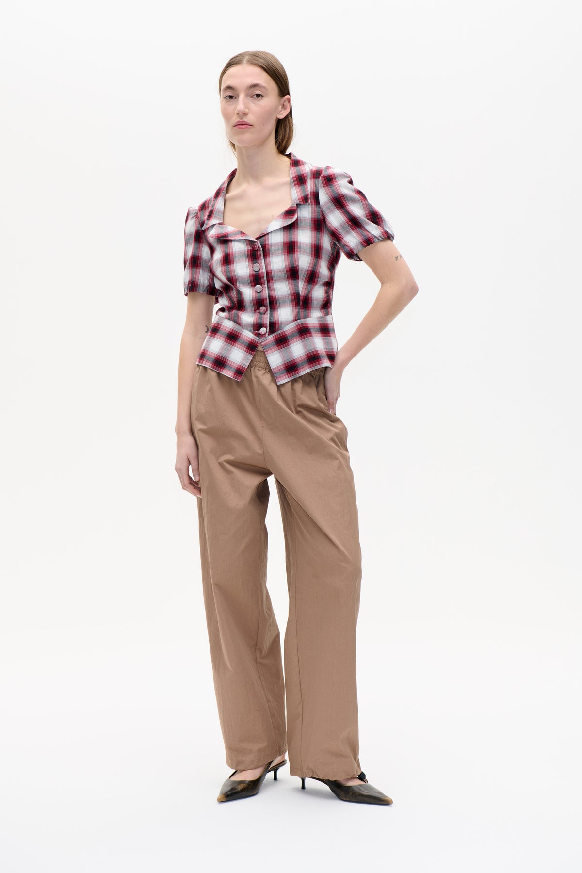 Mannequin portant un chemisier à carreaux rouge, noir et blanc, boutonné sur le devant et à manches bouffantes, avec un pantalon large couleur camel, vu de face.