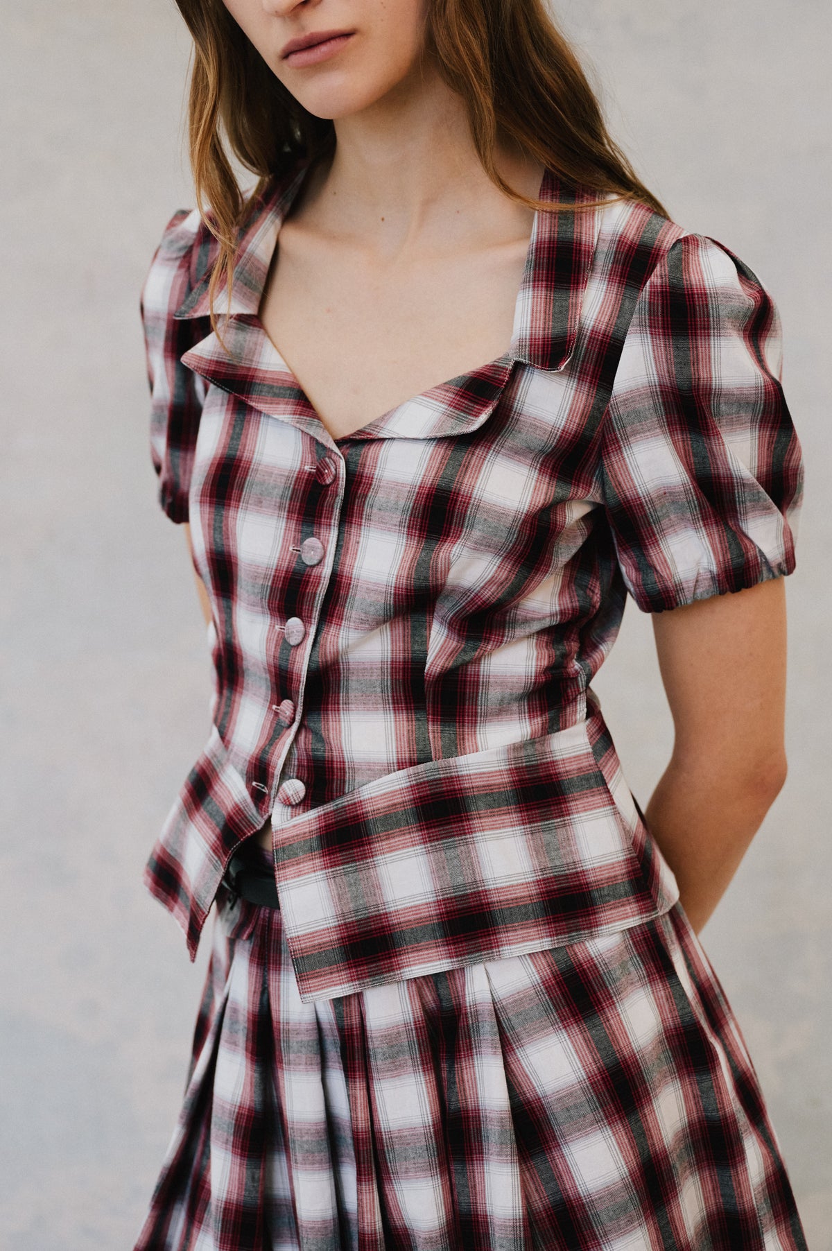 Portrait de campagne en gros plan d'un mannequin portant une robe à carreaux rouge, blanc et vert foncé, avec boutonnage sur le devant, manches bouffantes et ceinture assortie à la taille.