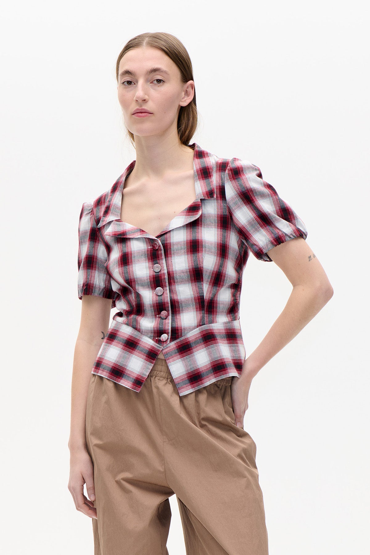 Mannequin portant un haut à carreaux rouge, bordeaux et blanc, boutonné sur le devant, à manches bouffantes et ourlet pointu, associé à un pantalon couleur camel.