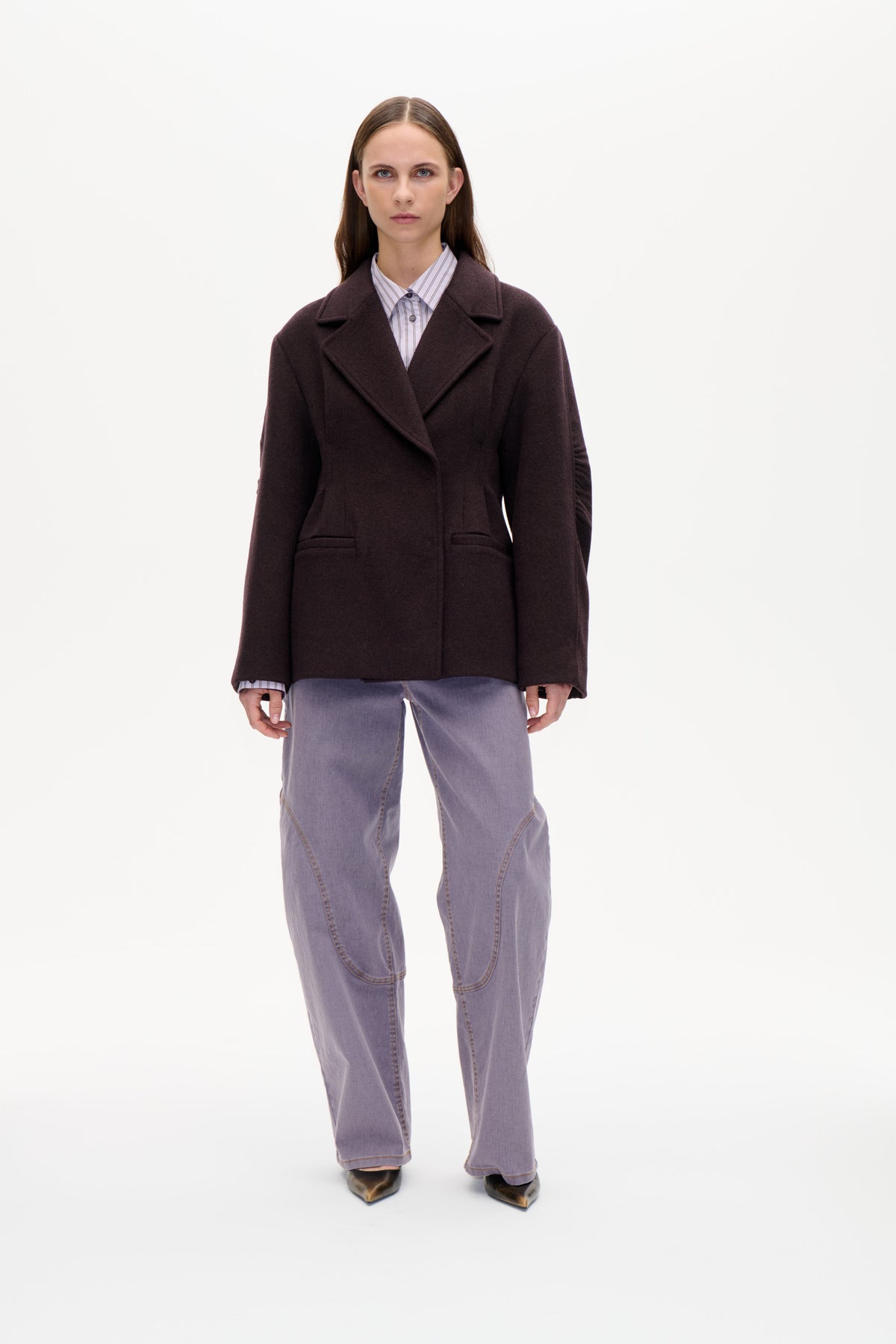 Front view of a model wearing a dark plum oversized notched-lapel coat over a striped shirt and light purple jeans.