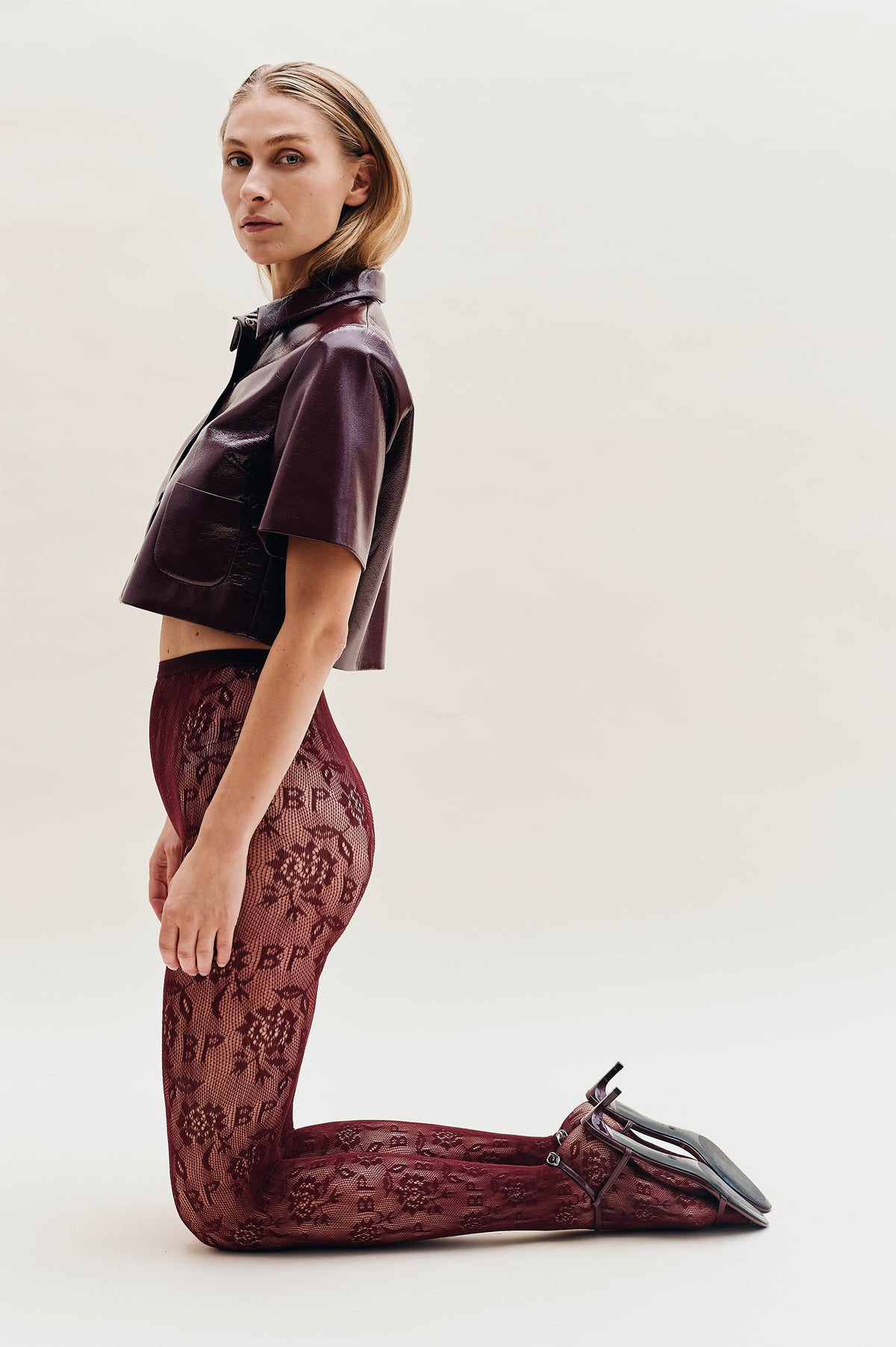 Model seated against a pale background in a glossy burgundy short-sleeved jacket and burgundy trousers with an intricate lace-like pattern.