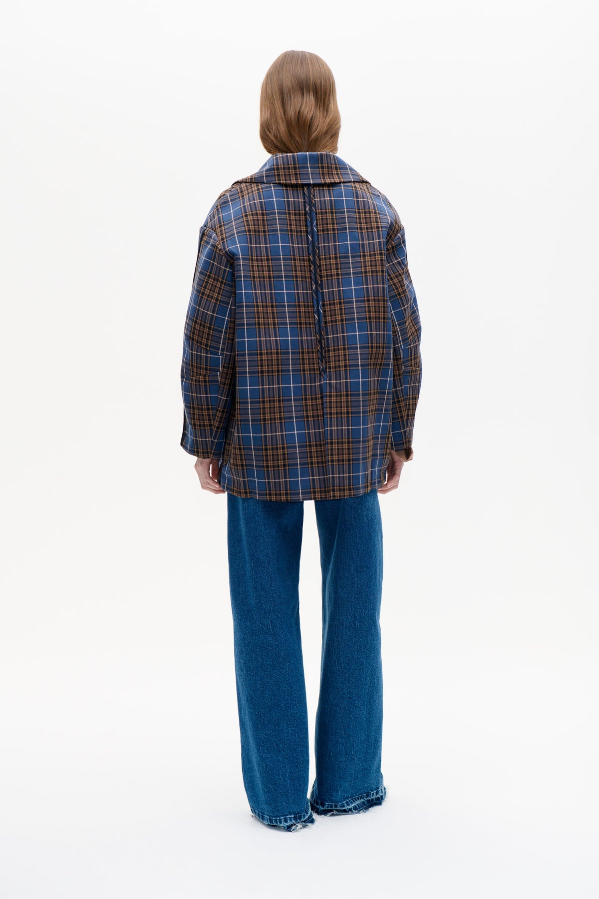 Back view of a model wearing a blue and brown plaid coat with a wide collar and central back seam, paired with wide-leg blue jeans.
