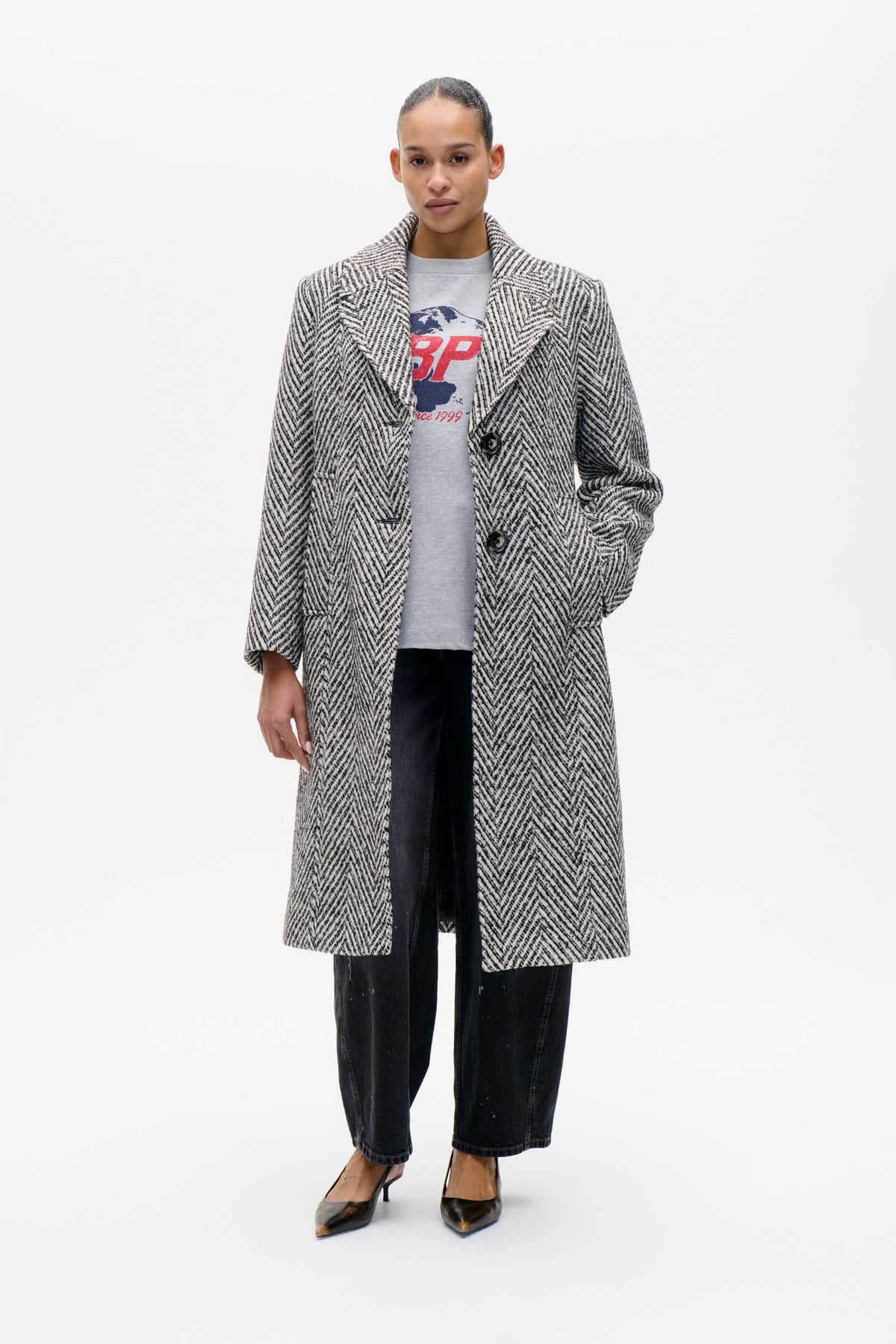 Model wearing a long black-and-cream herringbone coat with wide lapels and large buttons over a grey graphic T-shirt and dark jeans.