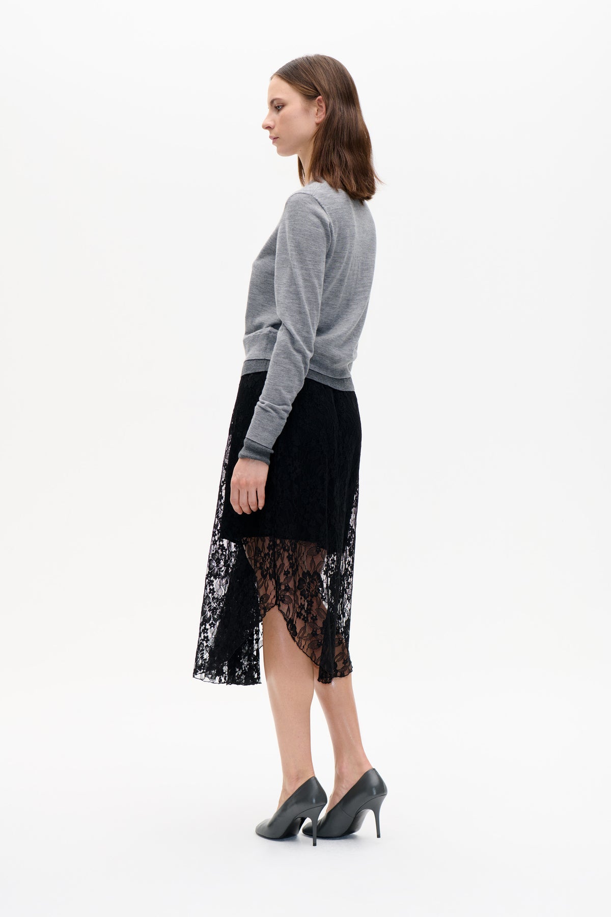 Side-profile fashion portrait of a model in a grey long-sleeved sweater, black lace skirt and black high heels against a white background.