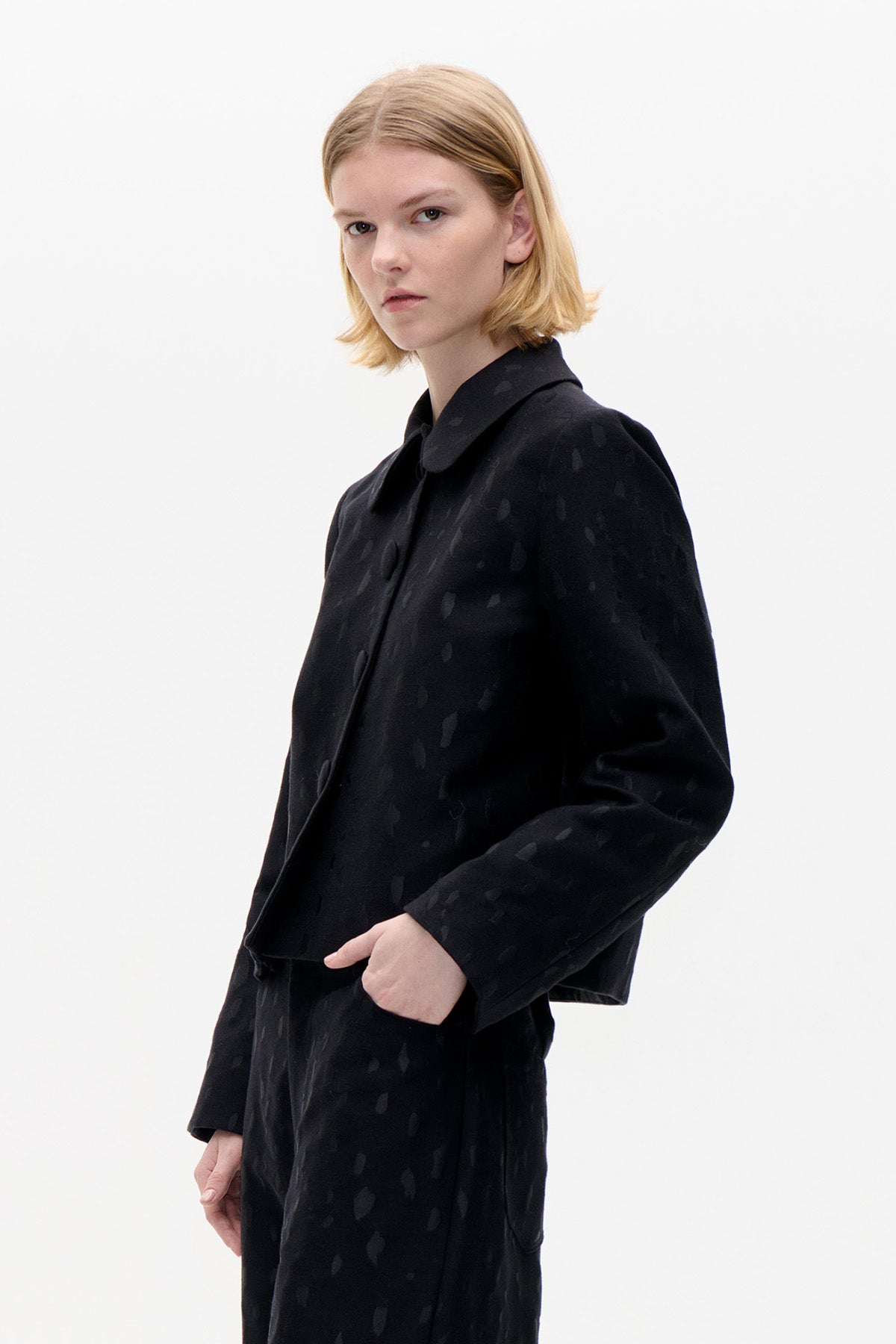 Model draagt een zwarte jas met een afgeronde kraag, bedekte knopen en een subtiel ton-sur-ton dessin, gecombineerd met een bijpassende broek.