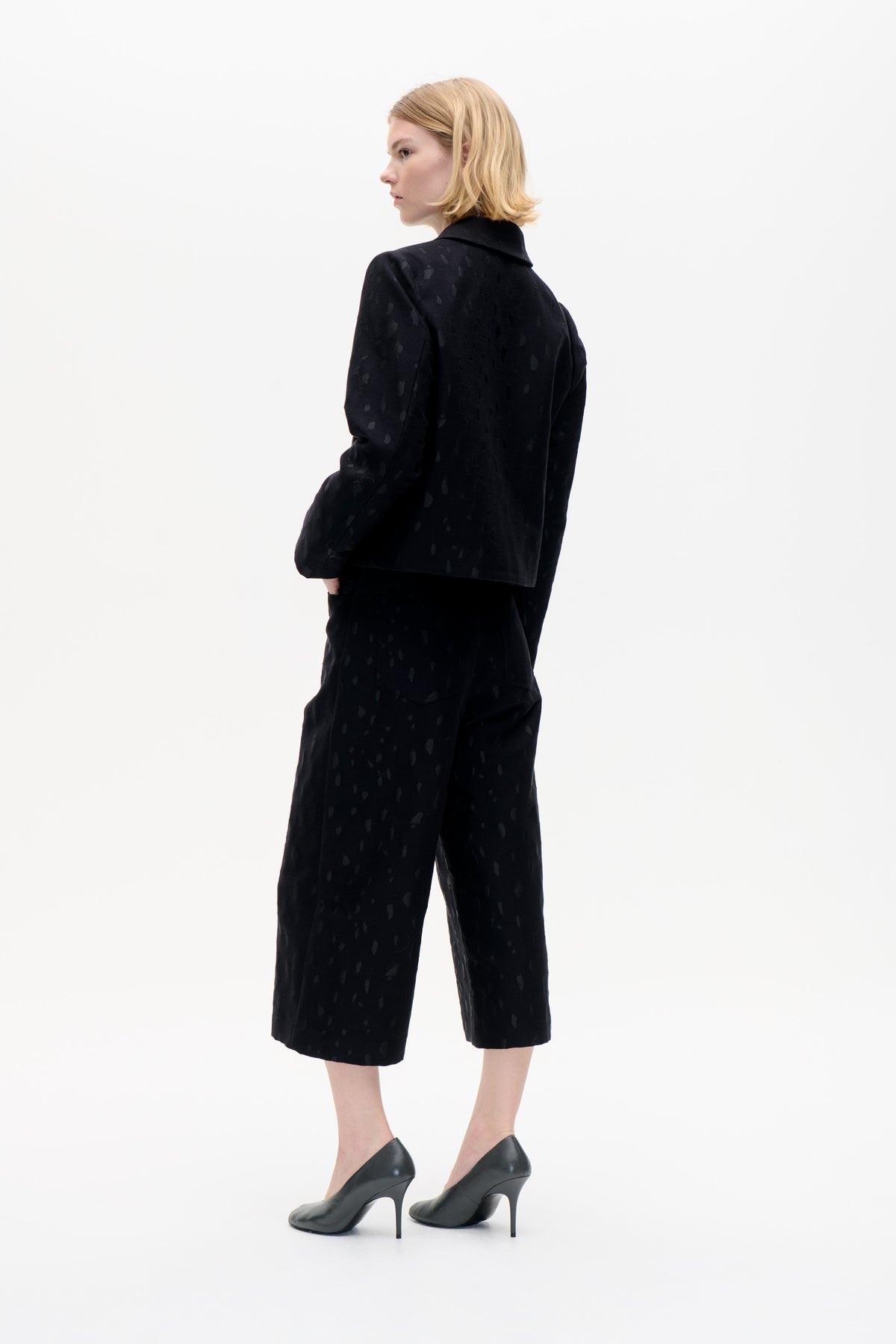 Model in a black collared jacket and matching cropped trousers with a subtle tonal abstract pattern, shown from the back and side.