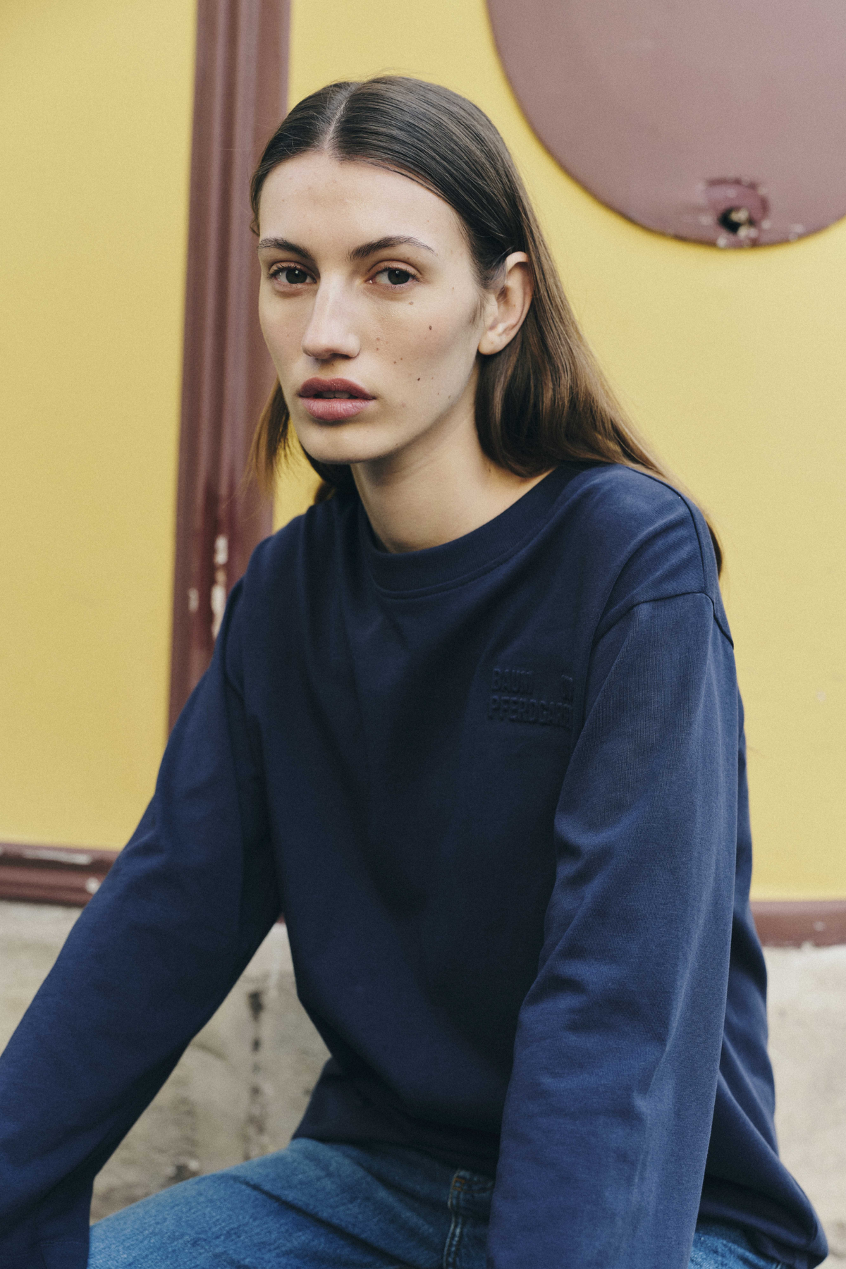 Model seated in a navy crew-neck sweatshirt with long sleeves and a subtle tonal chest detail, paired with blue jeans.