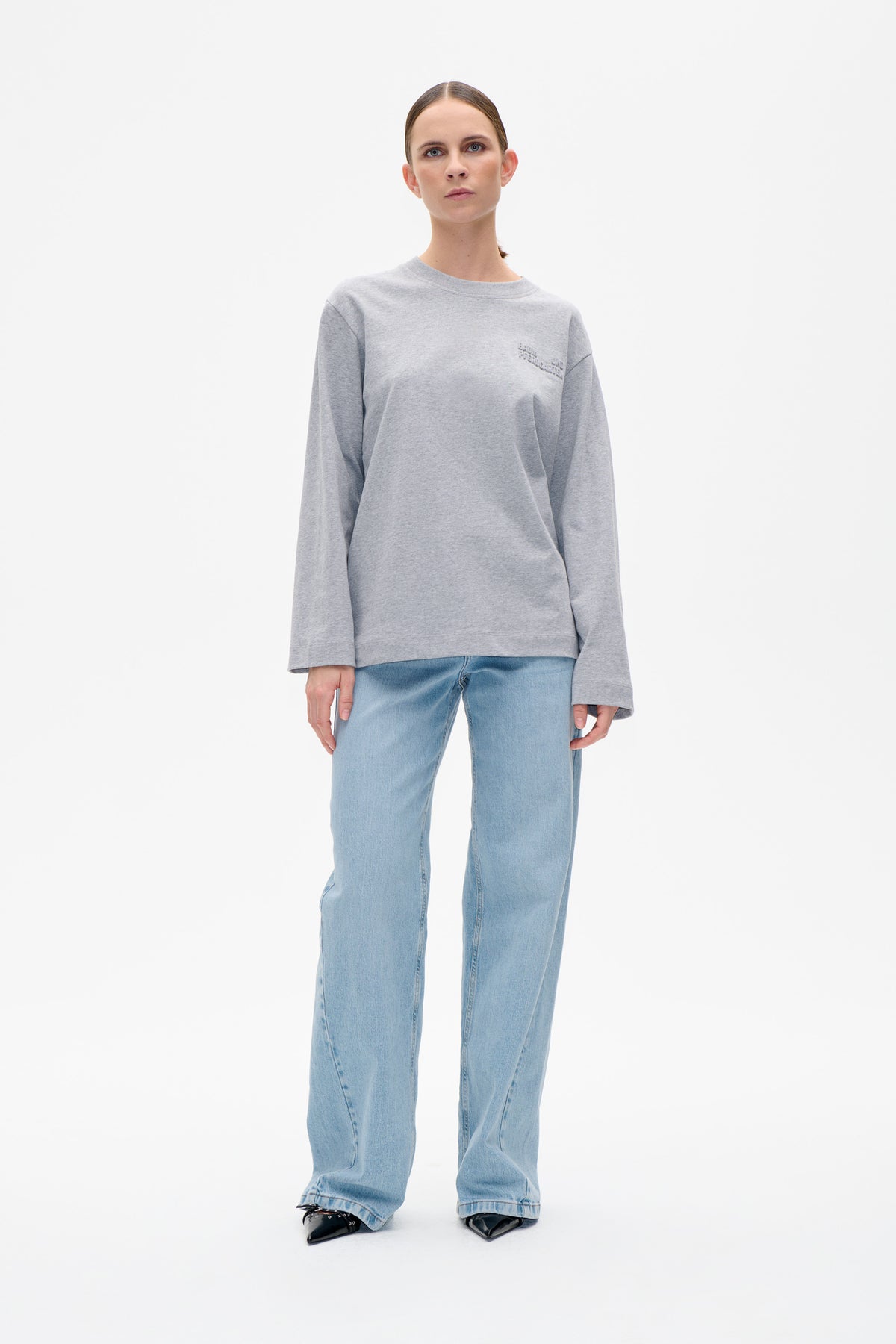 Front view of a model wearing a grey crewneck sweatshirt with tonal chest lettering and light-wash wide-leg jeans.