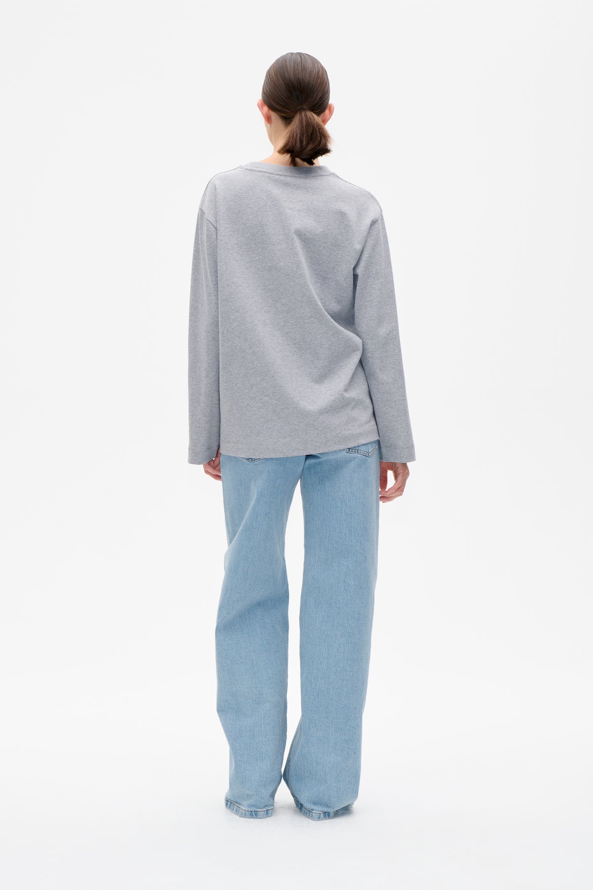 Back view of a model wearing a plain light grey long-sleeved sweatshirt with light blue wide-leg jeans.