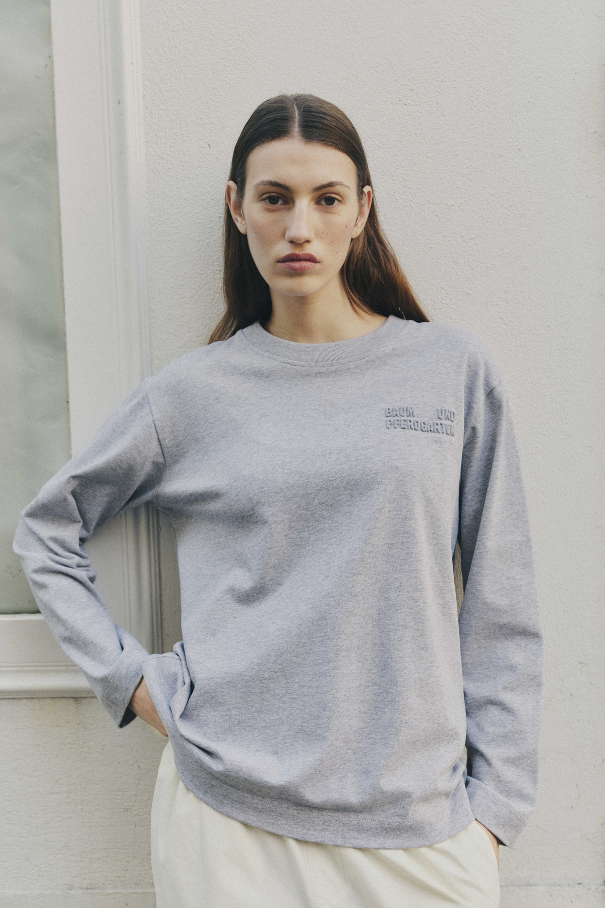 Front view of a model wearing a light grey crew-neck sweatshirt with subtle tonal chest lettering and cream trousers.