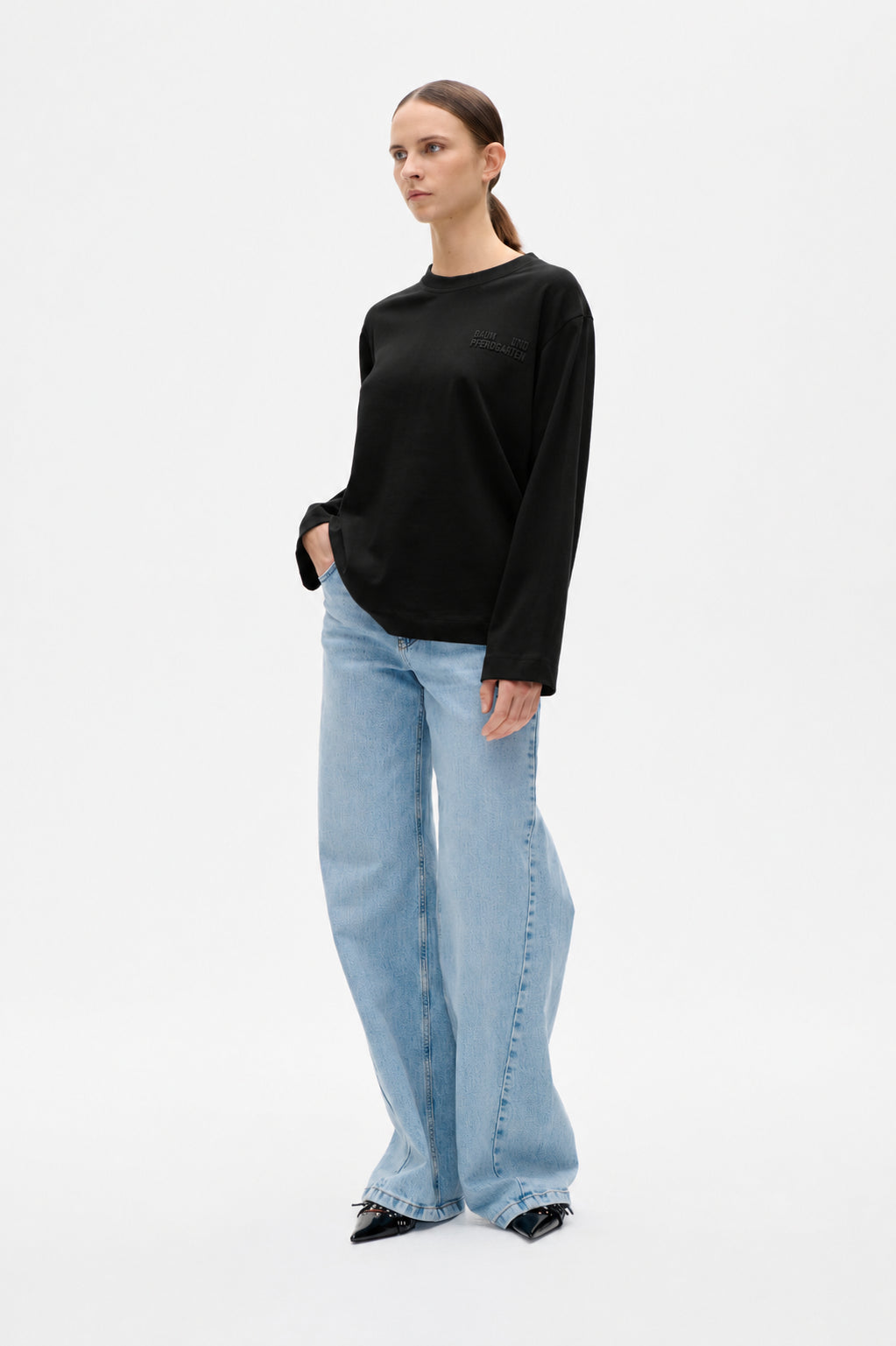 Model wearing a black long-sleeve crewneck top with a small tonal chest logo and light blue wide-leg jeans.