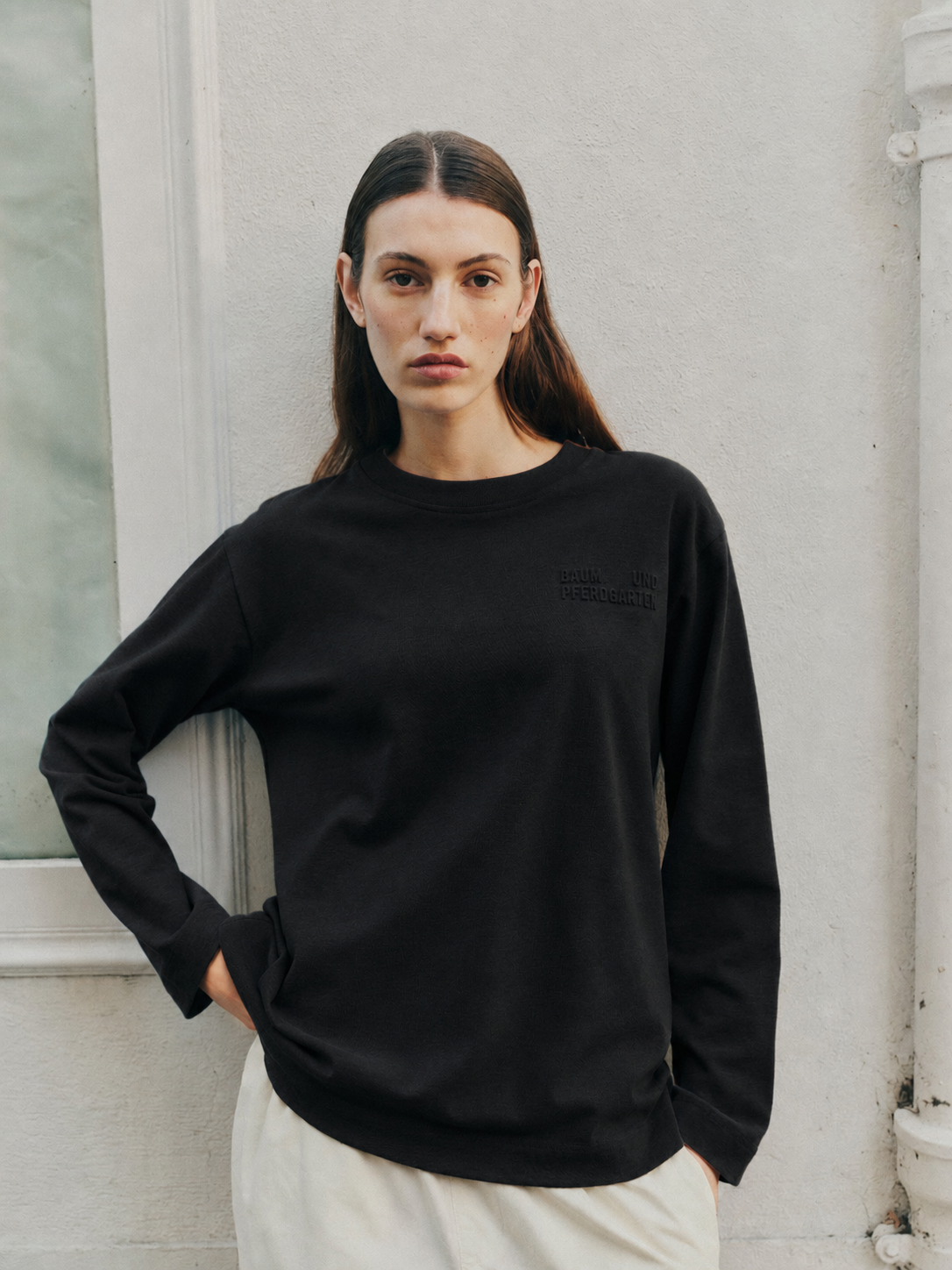 Model wearing a black long-sleeved crewneck sweatshirt with subtle embossed chest lettering and white trousers.