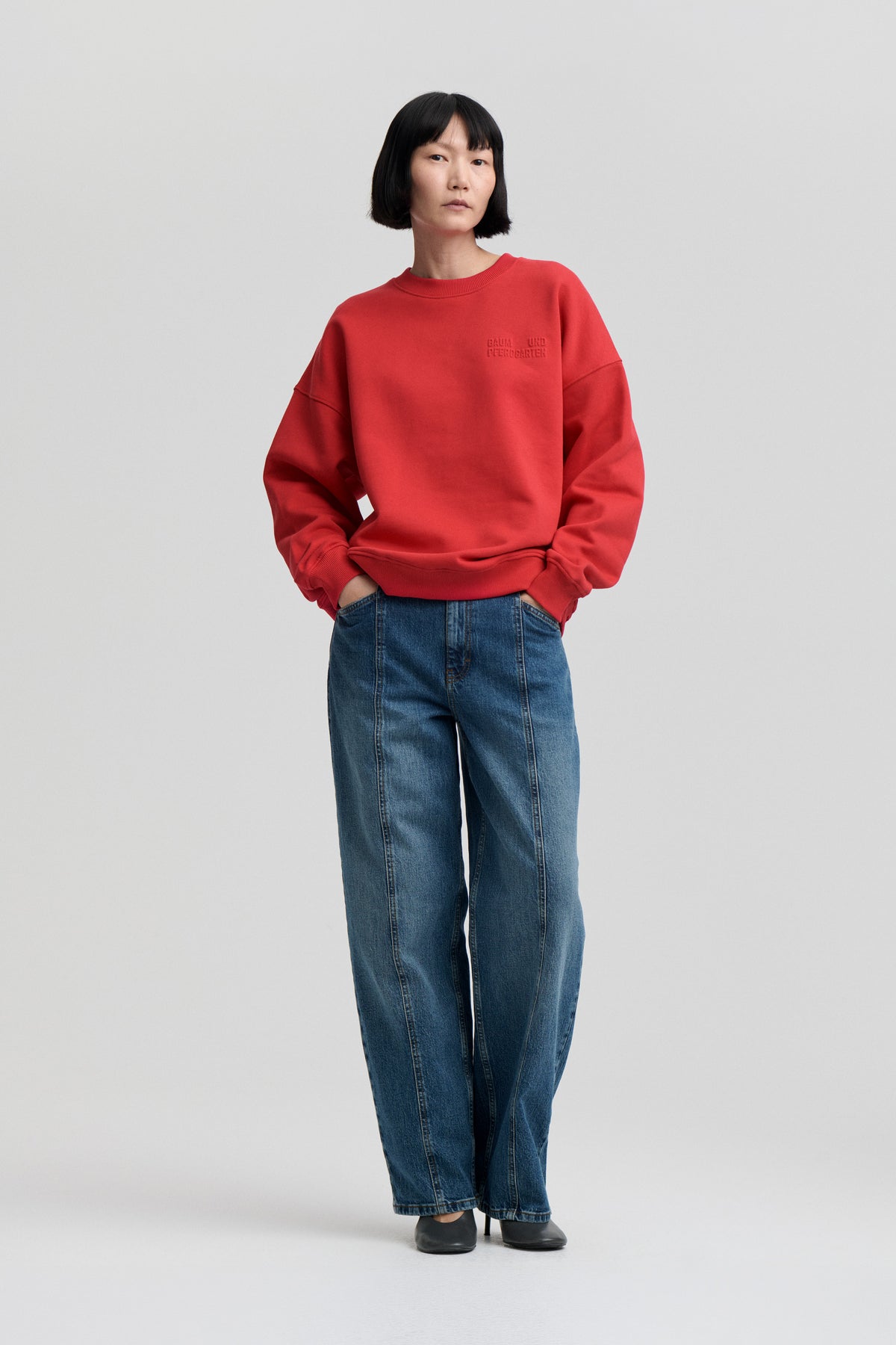 Model wearing a bright red oversized crewneck sweatshirt with tonal chest lettering and blue wide-leg jeans, shown from the front.
