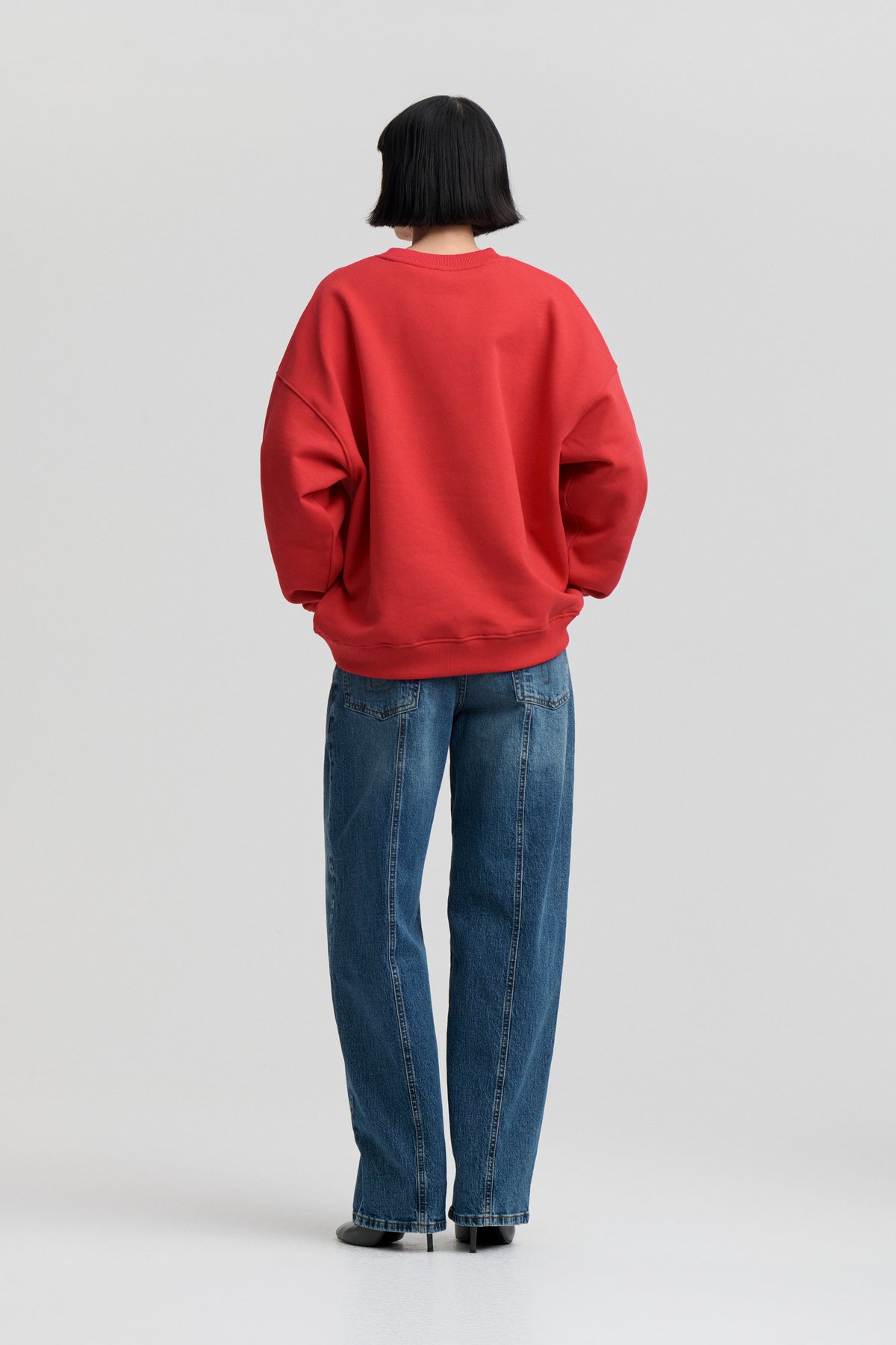 Back view of a model wearing an oversized red sweatshirt with ribbed cuffs and hem, paired with blue jeans.