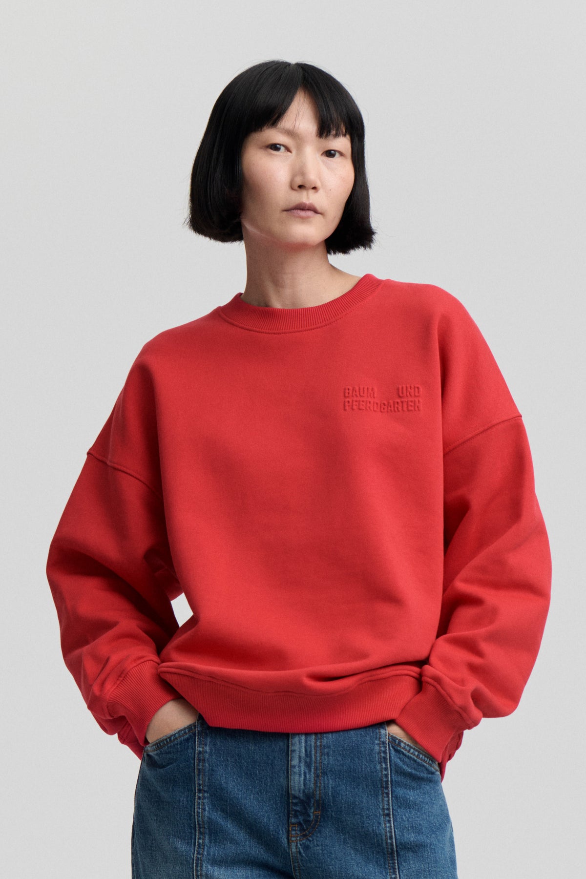 Model wearing a bright red crew-neck sweatshirt with tonal chest lettering and ribbed trims, paired with blue jeans.