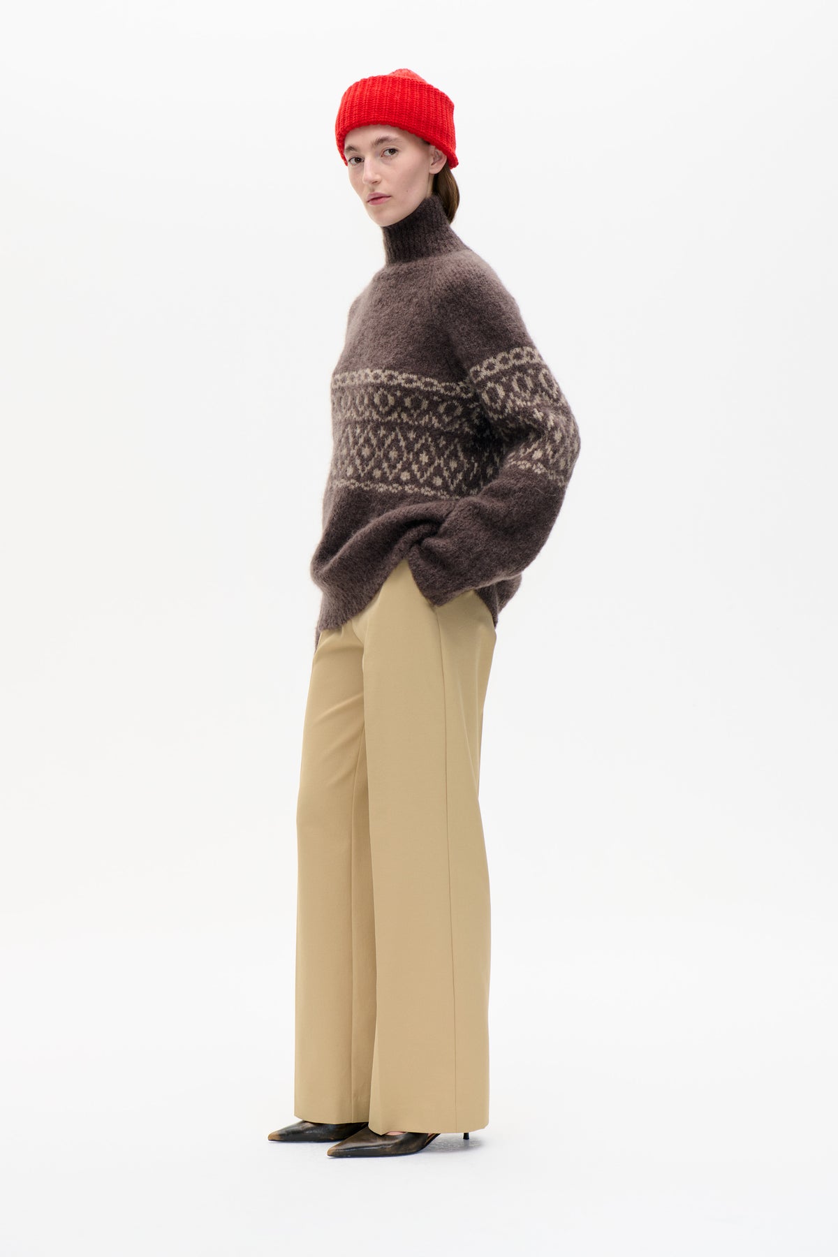 Model wearing a dark brown high-neck sweater with cream geometric bands, a bright red knitted beanie and beige wide-leg trousers.