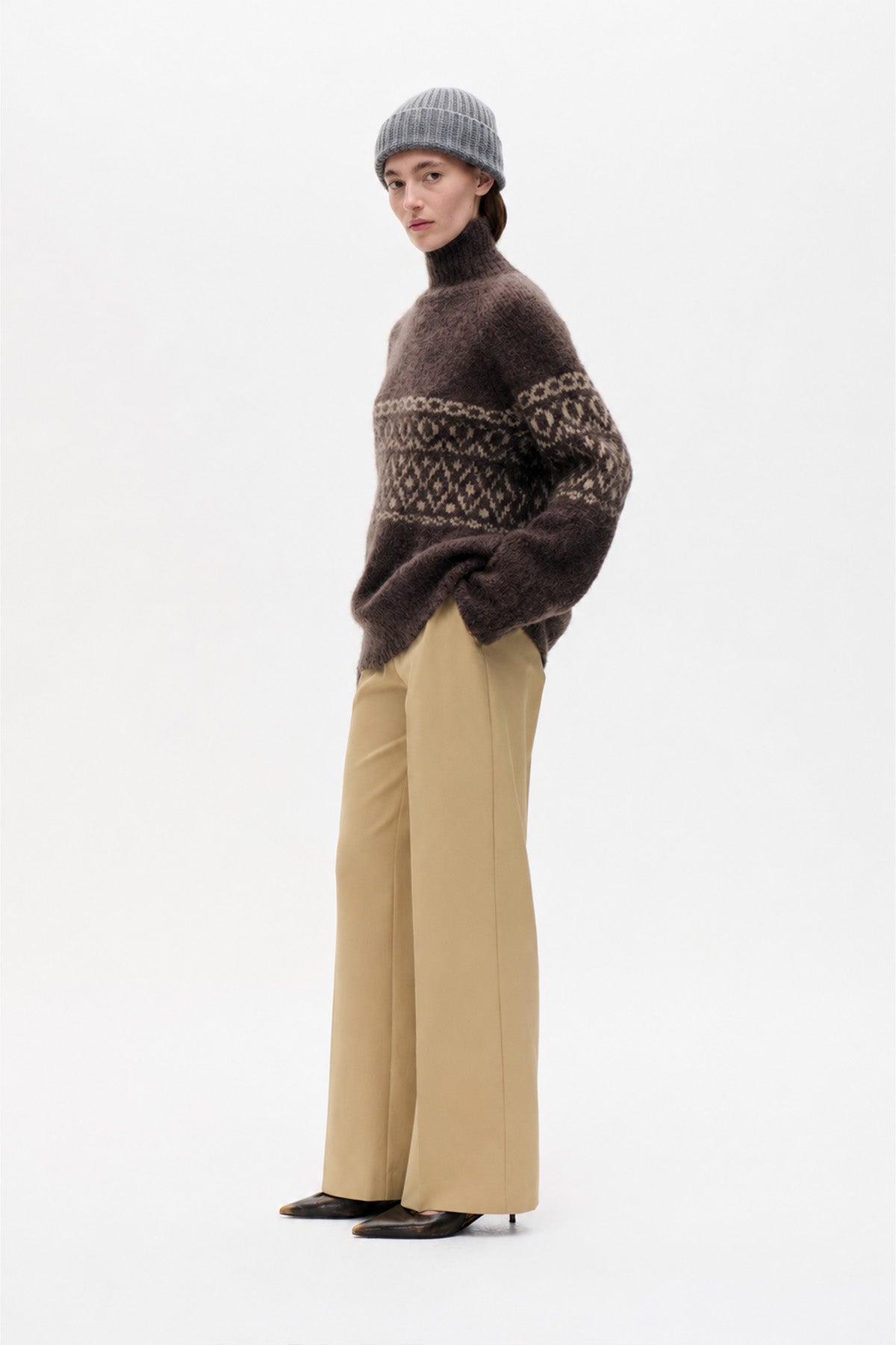 Model wearing a dark brown high-neck sweater with a cream geometric pattern, wide-leg beige trousers and a grey knit beanie.