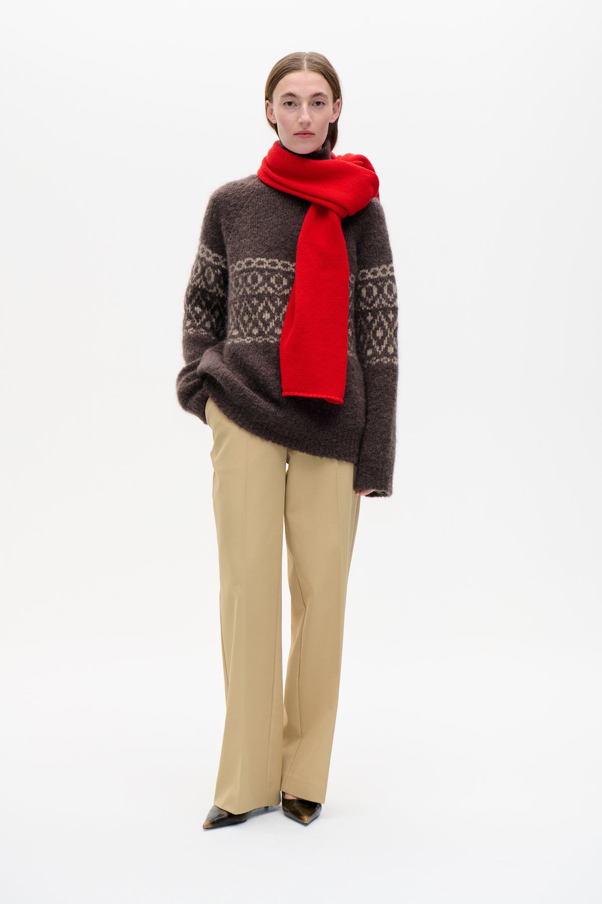 Front view of a model wearing a dark brown patterned knit sweater, bright red scarf and beige wide-leg trousers against a white background.