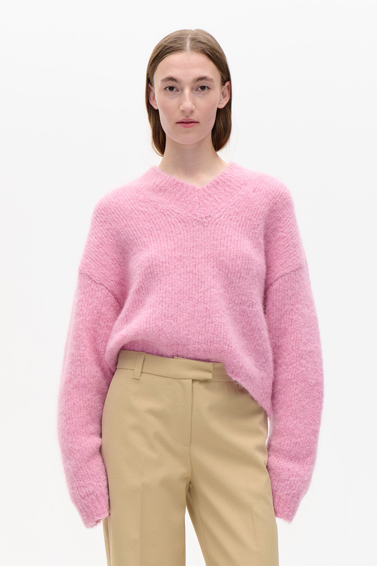 Front view of a model wearing a fuzzy pink V-neck sweater with dropped shoulders and beige trousers.