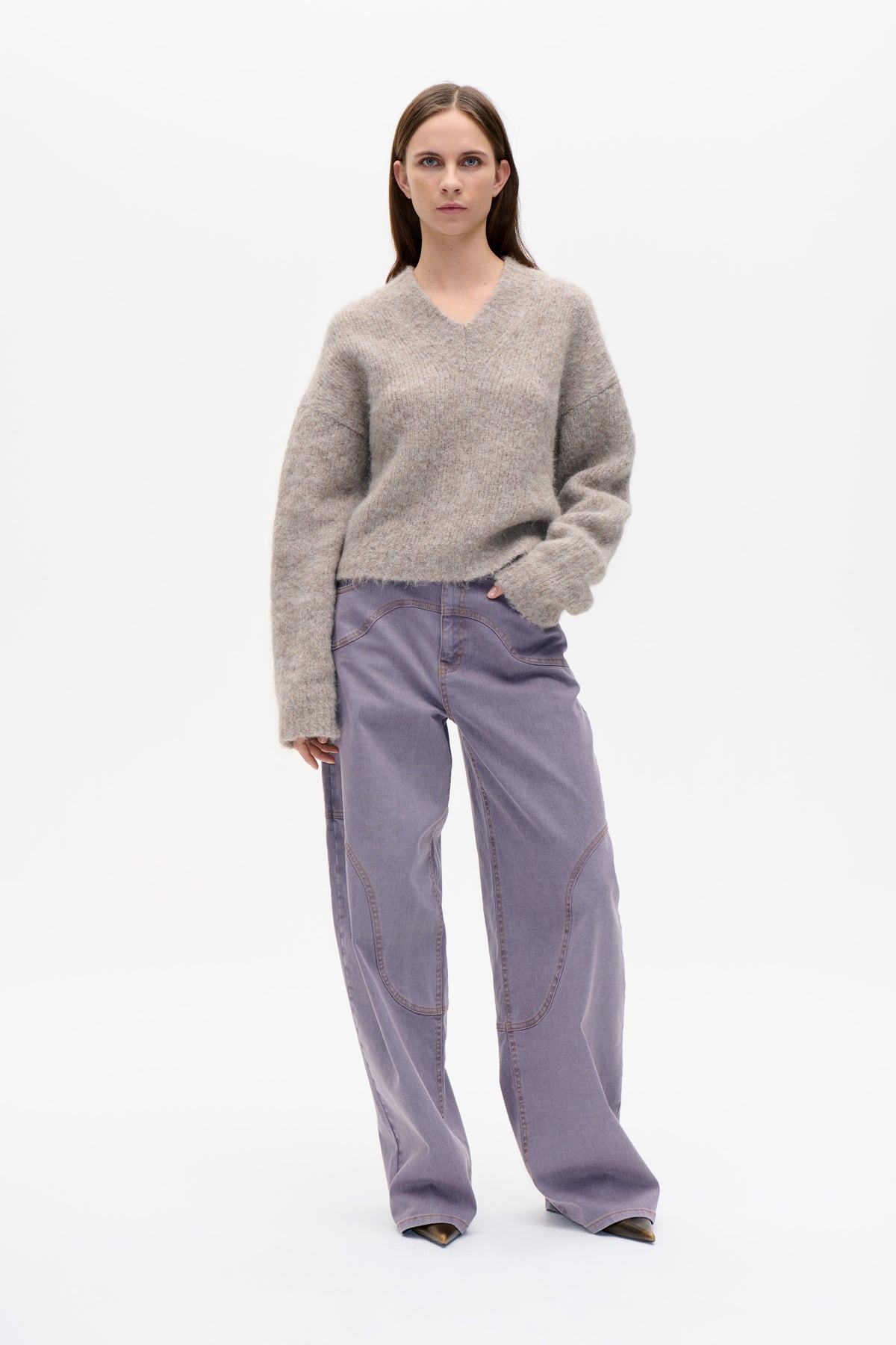 Model in a loose taupe marled V-neck sweater and lilac wide-leg jeans with curved contrast-stitched panels against a white studio background.
