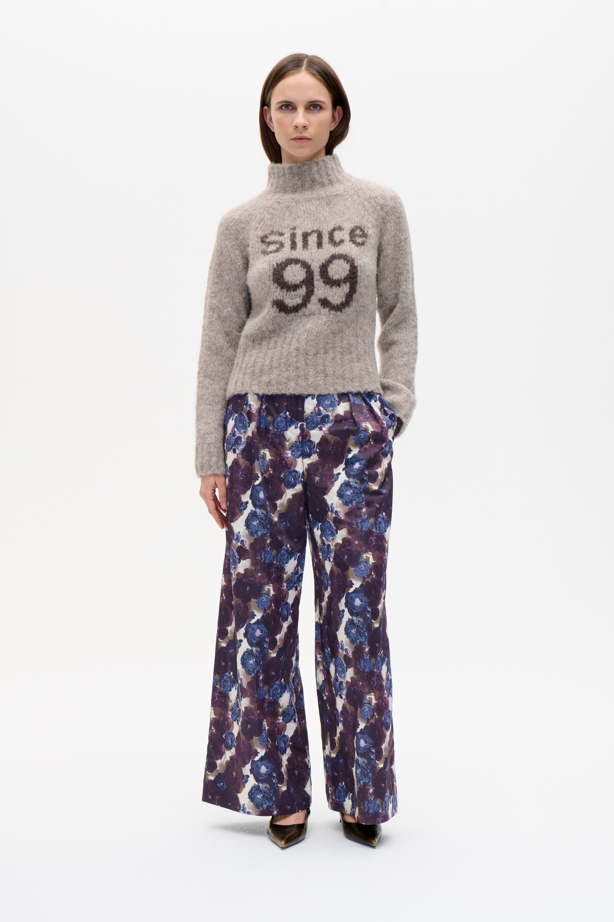Front-facing model wearing a taupe high-neck sweater with dark lettering and a smiley motif, paired with blue and purple floral trousers.