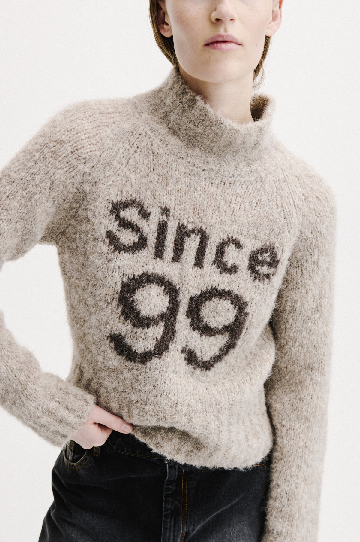 Close-up campaign view of a model wearing a fuzzy beige sweater with a bold black abstract pattern and dark jeans.